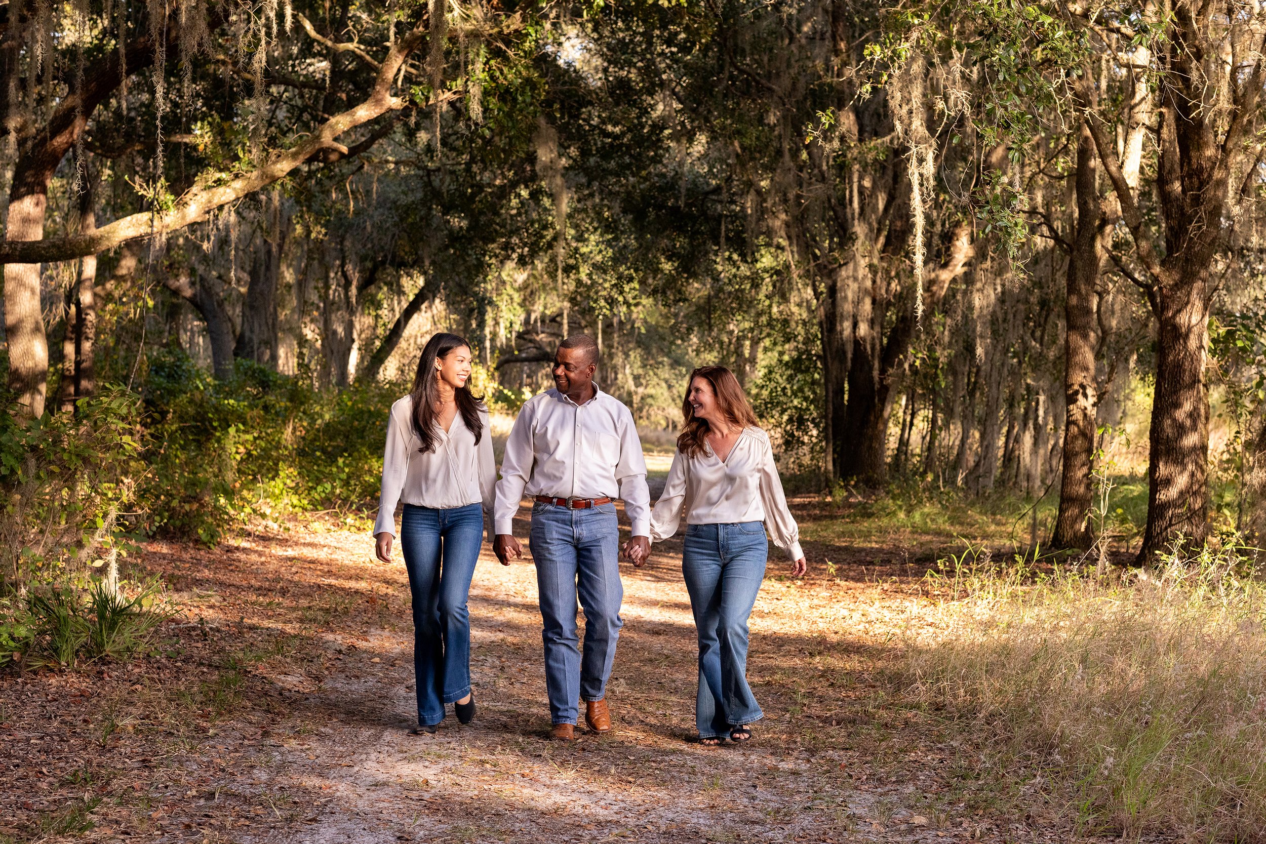 Lifestyle family photography of three people walking hand in hand along a wooded path during golden hour