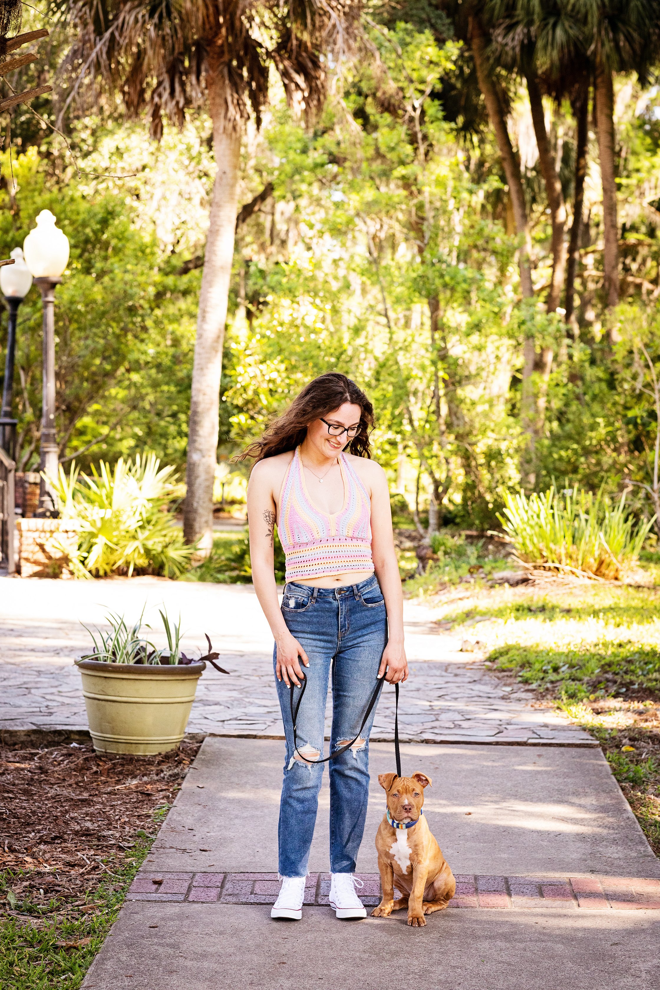 Lifestyle portrait of a woman walking her dog in a sunlit park setting, looking down at a brown dog sitting beside her. She is wearing a colorful, sleeveless crop top, ripped jeans, and white sneakers. 