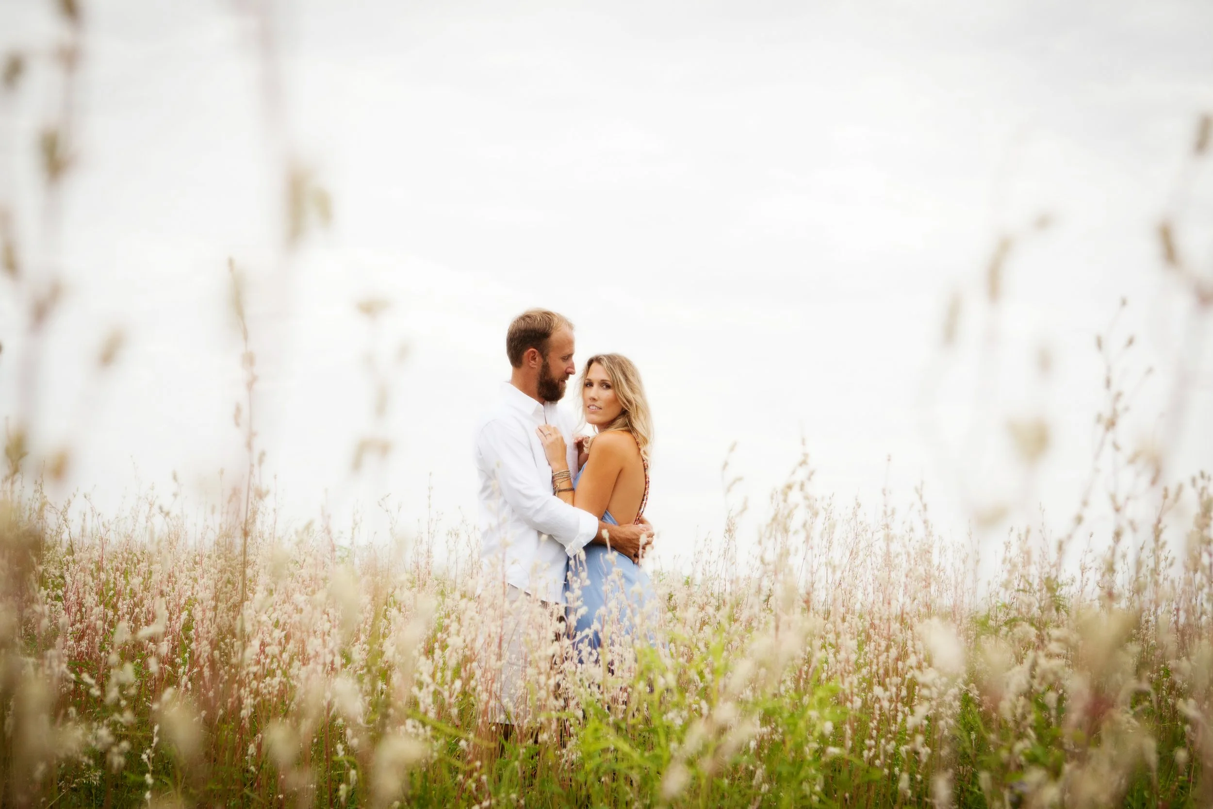 Lifestyle couple photography of a man and woman standing close together in a field of flowers, with a bright, overcast sky in the background.