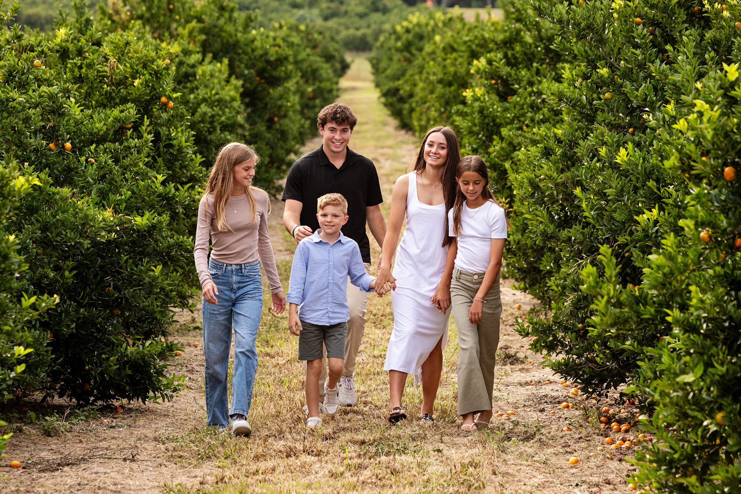Lifestyle group photography of a group of six people, including children of various ages, walking through a Florida orange grove holding hands and smiling.