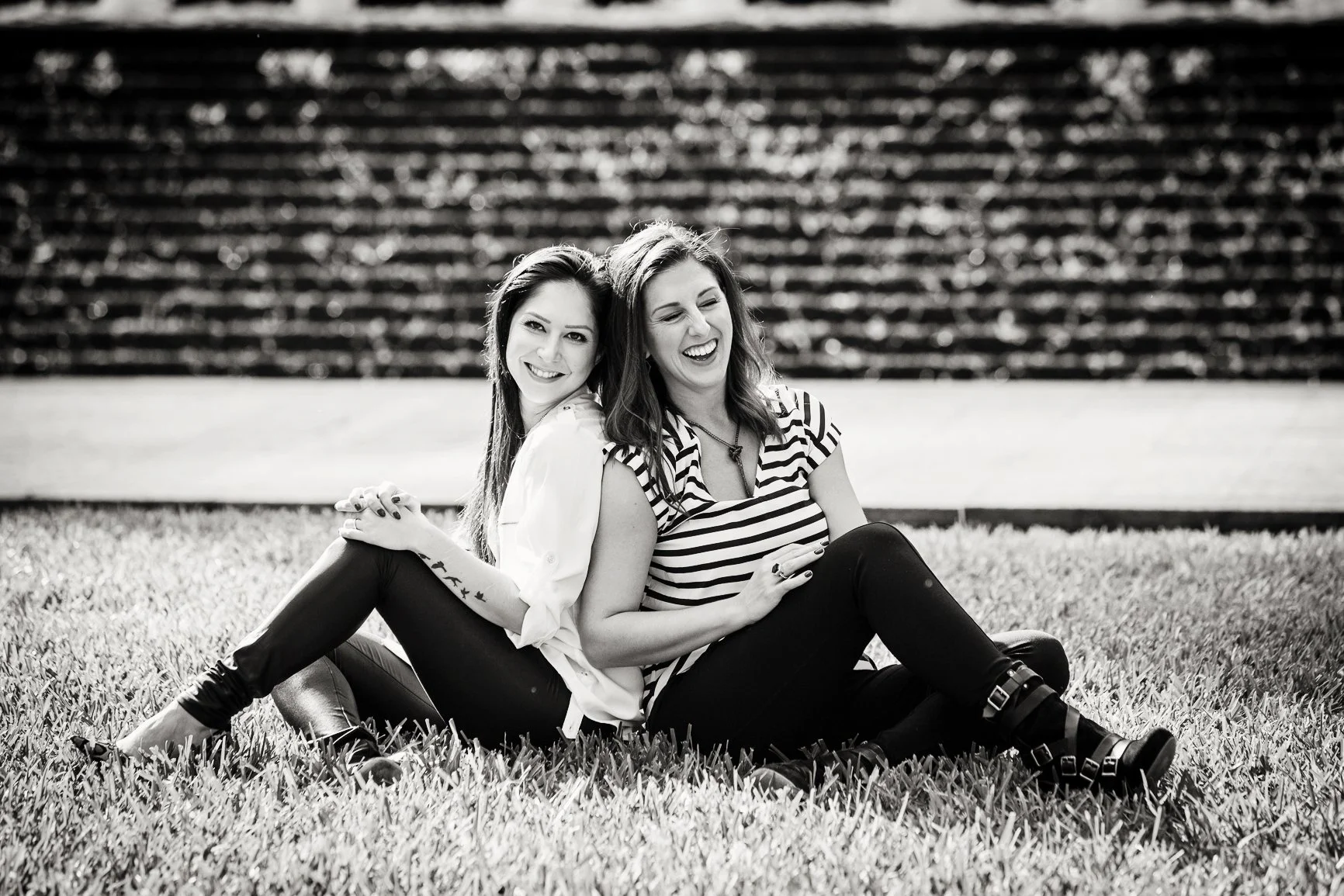Lifestyle and brand portrait photography showing two women sitting back-to-back on grass, laughing together