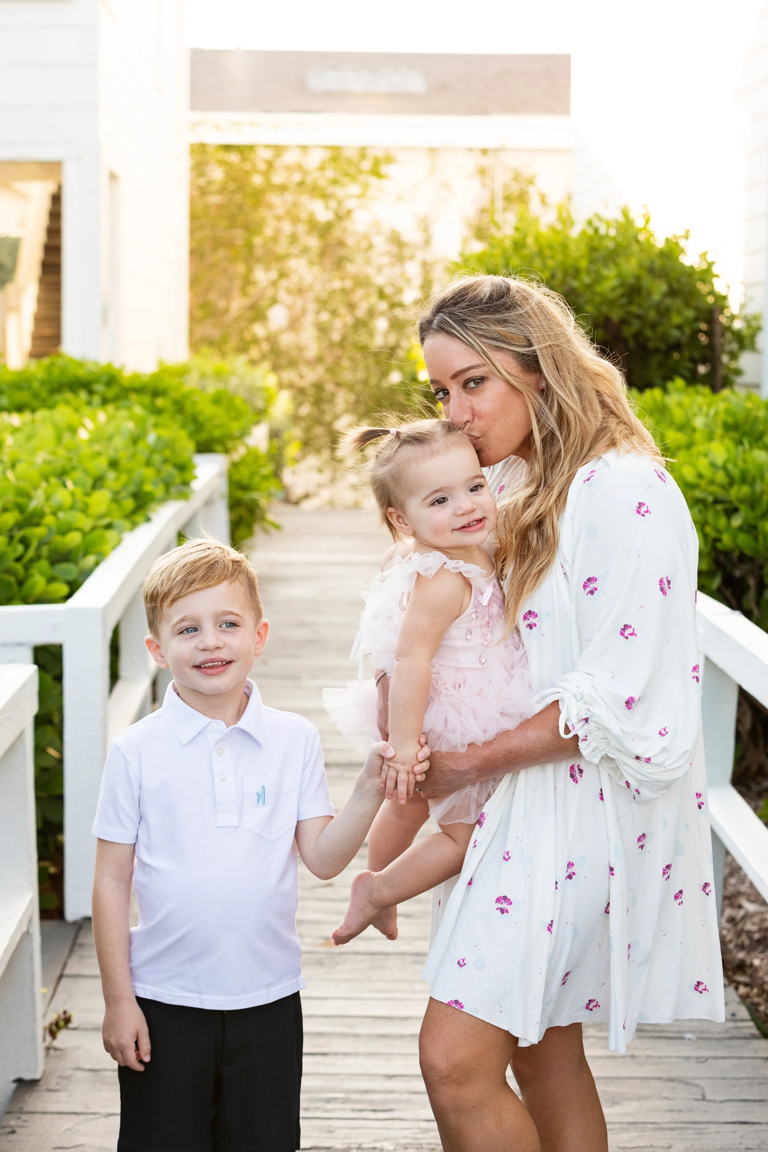 Lifestyle family portrait of a mother kissing her daughter while her son holds her hand on a bridge at sunset.