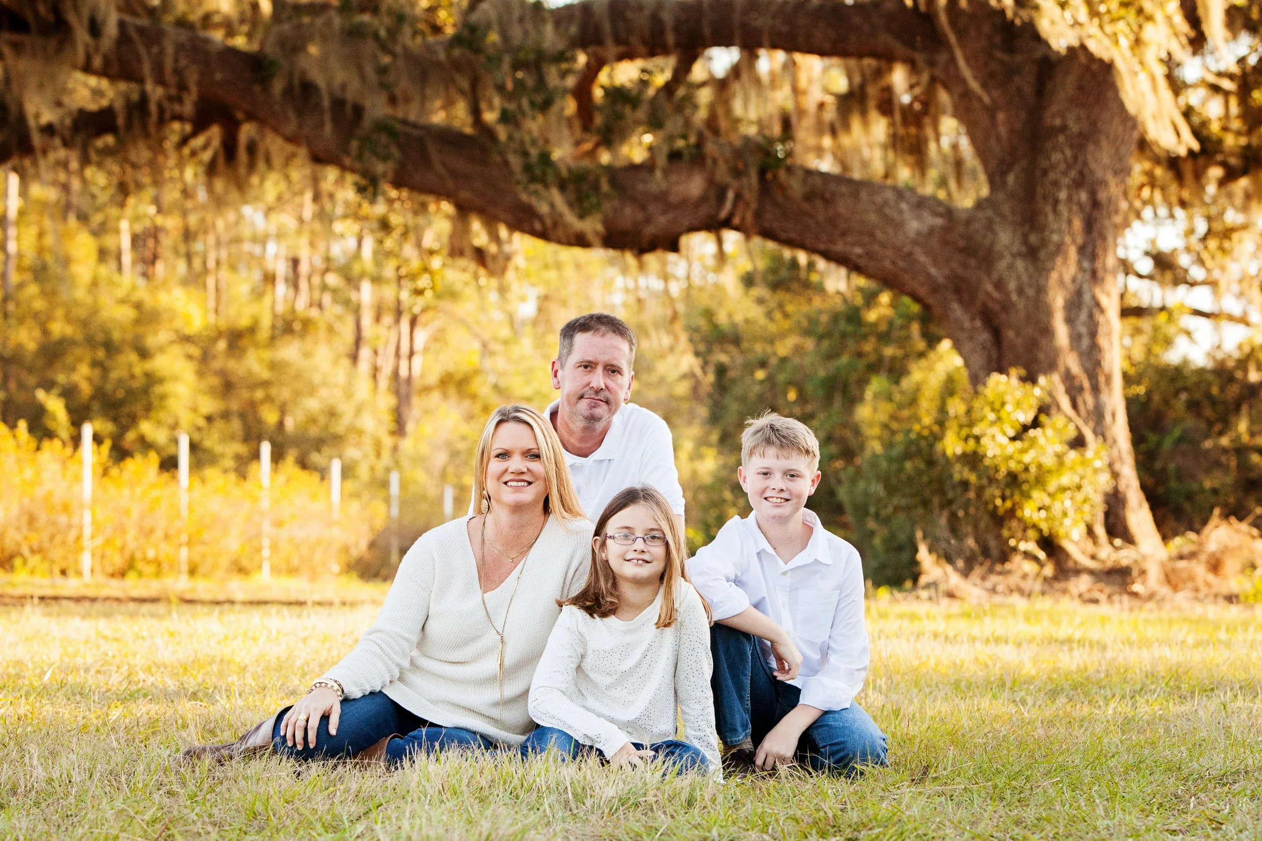 Outdoor family photography of a family of four sitting beneath a tree with fall foliage at sunset.
