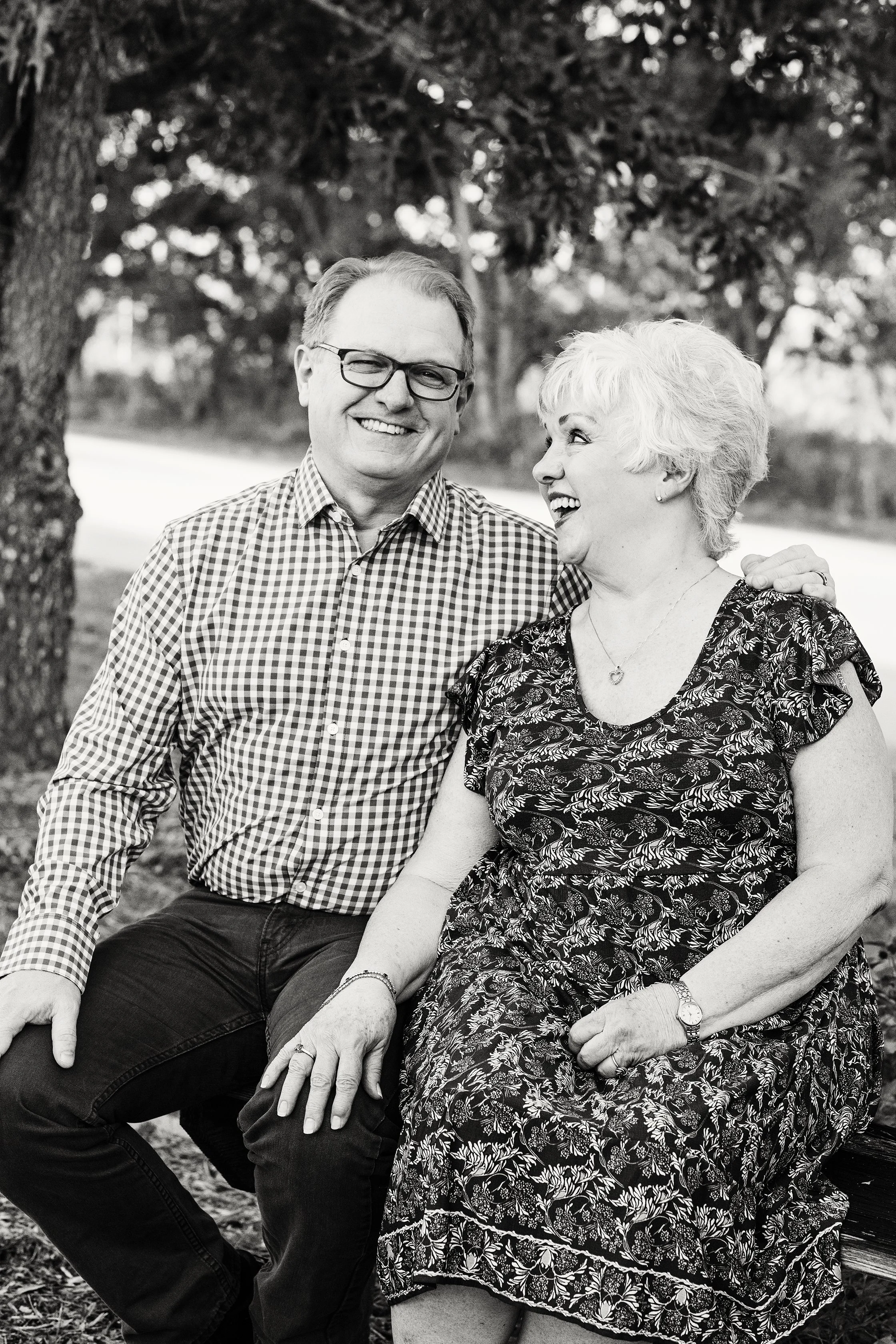 Black and white lifestyle couple portrait of a man and woman sitting together outdoors, laughing beneath trees in their golden years.