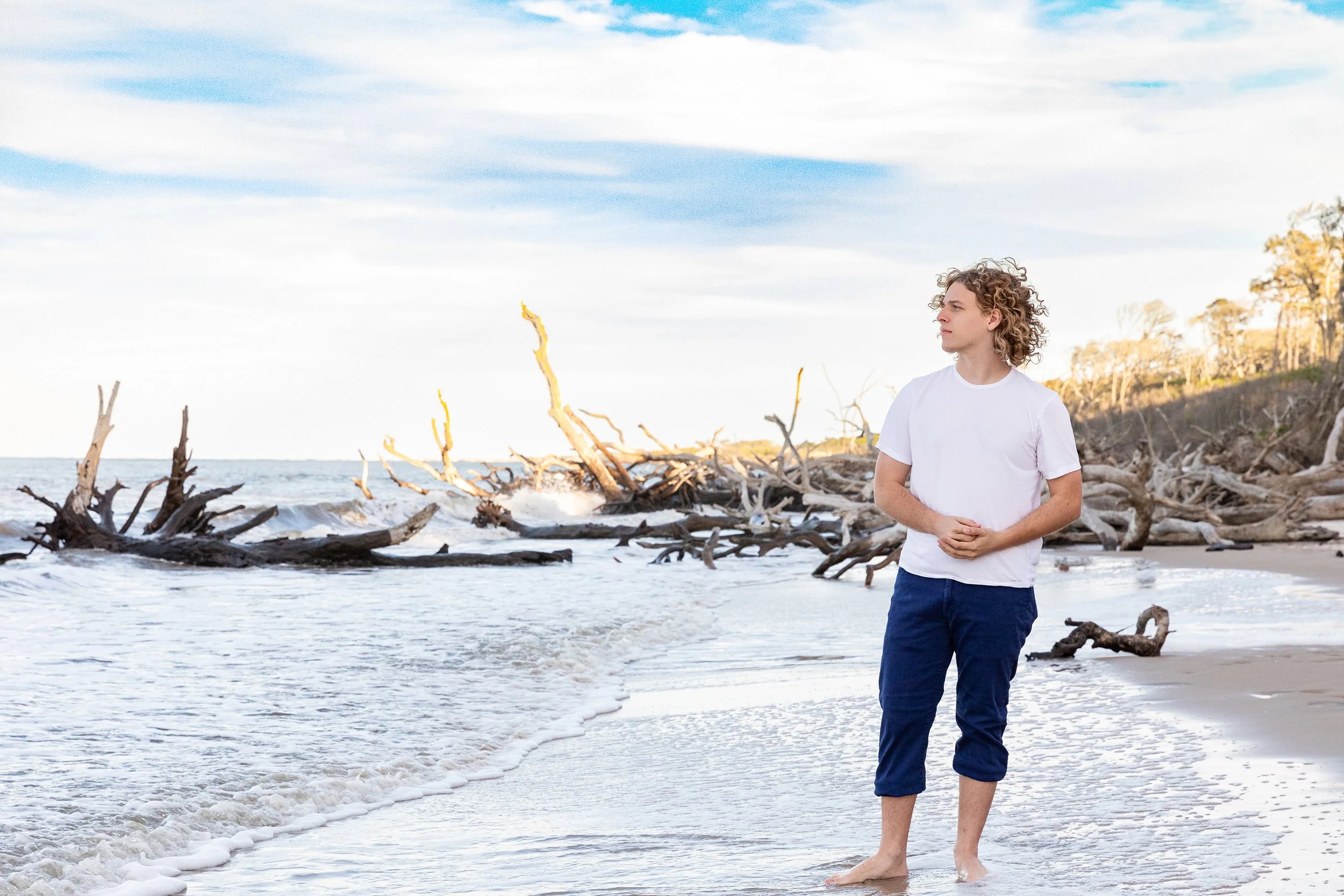 Floriday lifestyle senior portrait of a young man standing near driftwood and trees along the shoreline, photographed by Lindsay Parks Photography