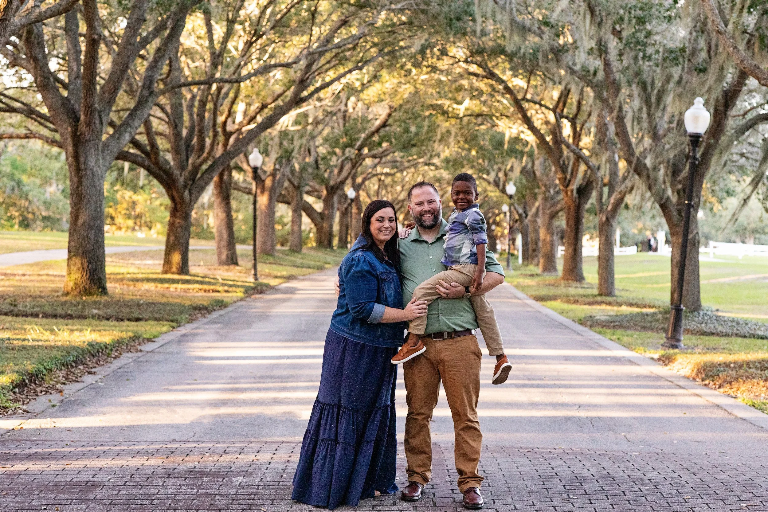 Outdoor family portrait photography of a family of three walking at sunset under Florida Spanish moss trees, photographed by Lindsay Parks Photography.