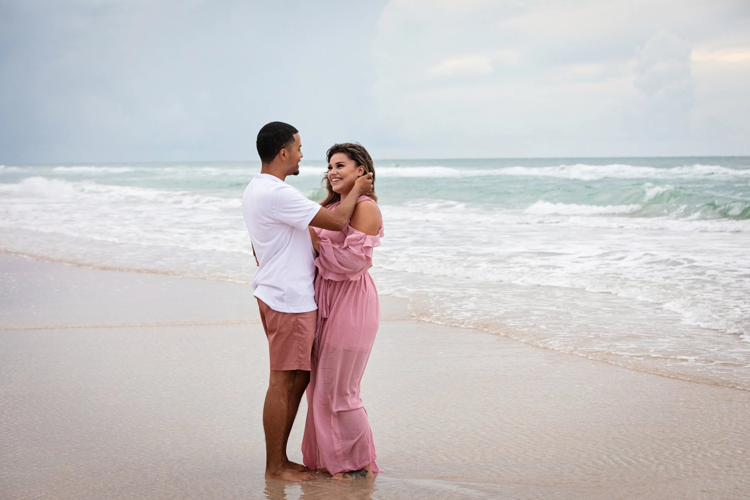 Beach couple photography of a couple standing together in shallow water smiling, smiling and looking at each other, with the ocean and cloudy sky in the background.