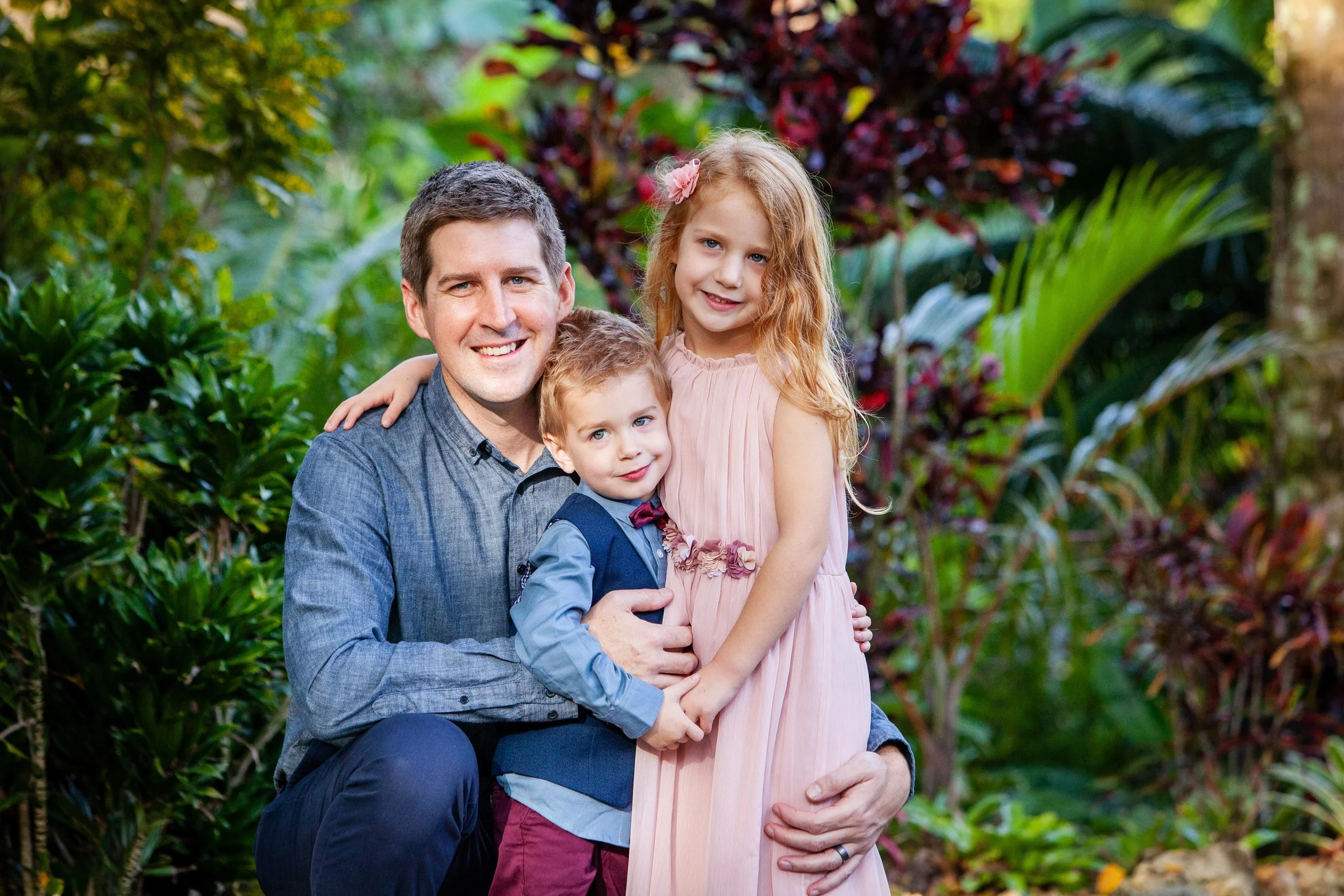 Lifestyle family photography of a father kneeling with his children in a lush garden The children are hugging the man and each other, surrounded by green tropical plants.