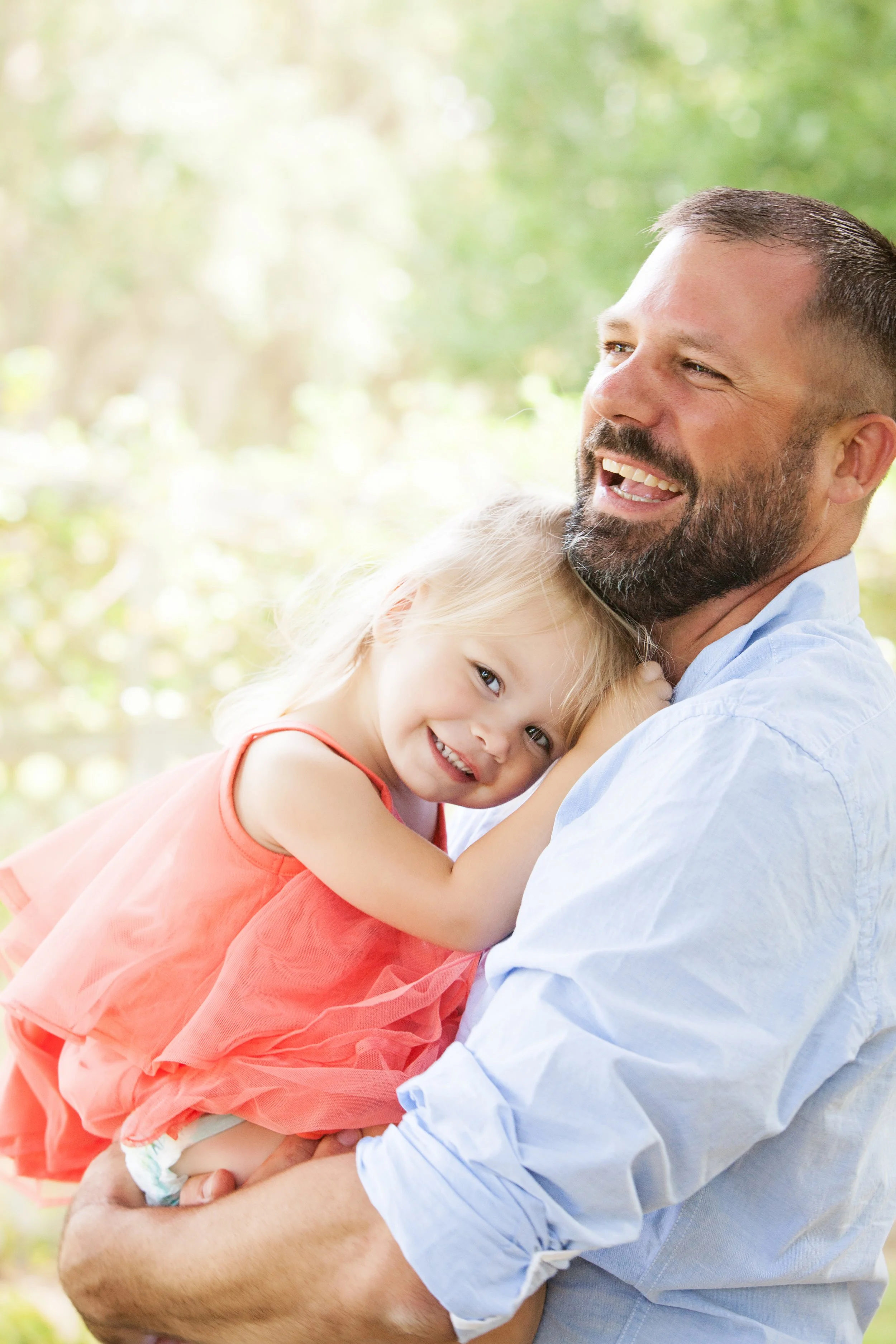 Lifestyle family portrait of a father holding his daughter and smiling beneath trees