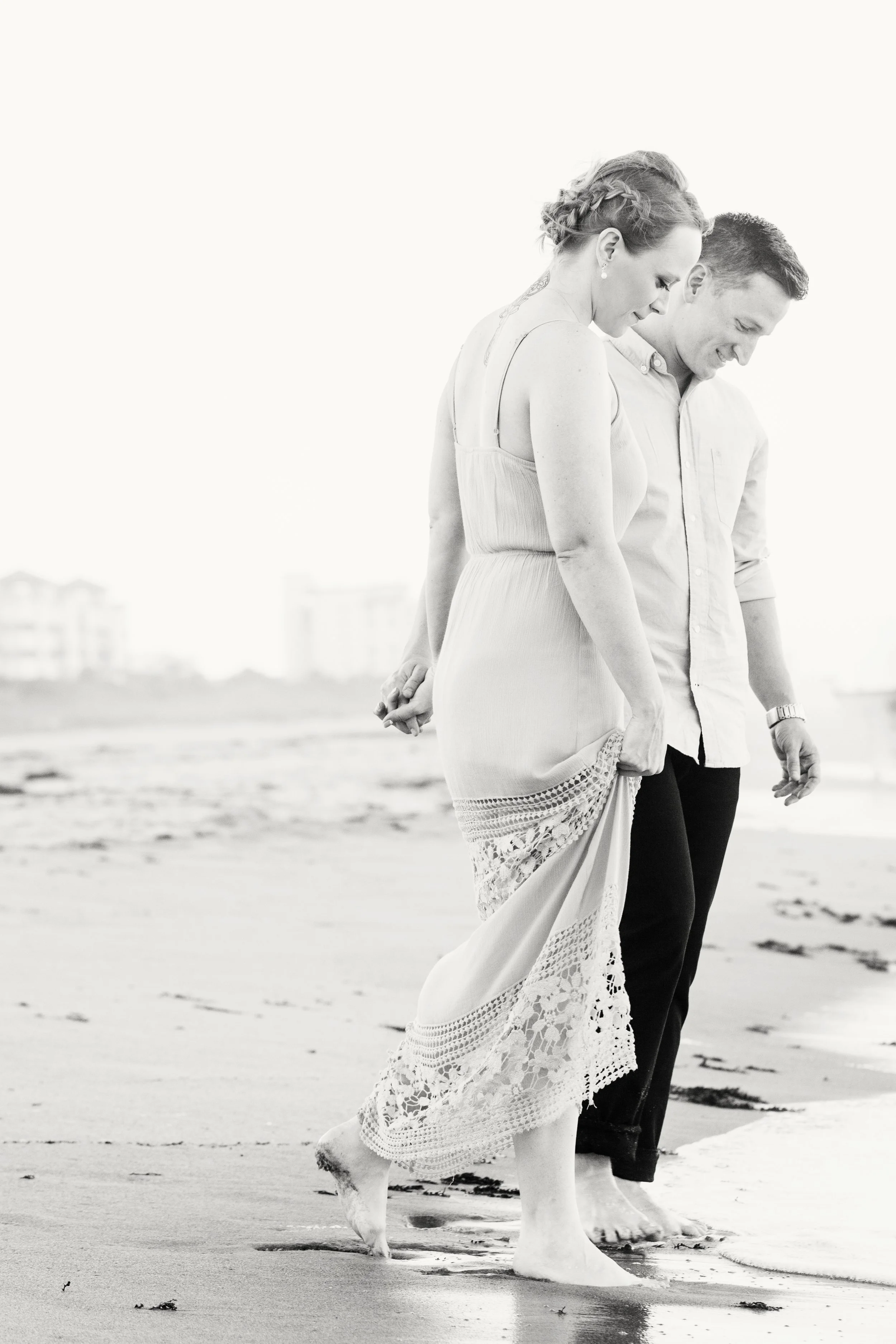 Black-and-white beach couple photography of a couple walking barefoot along the Florida shoreline, holding hands behind their backs.