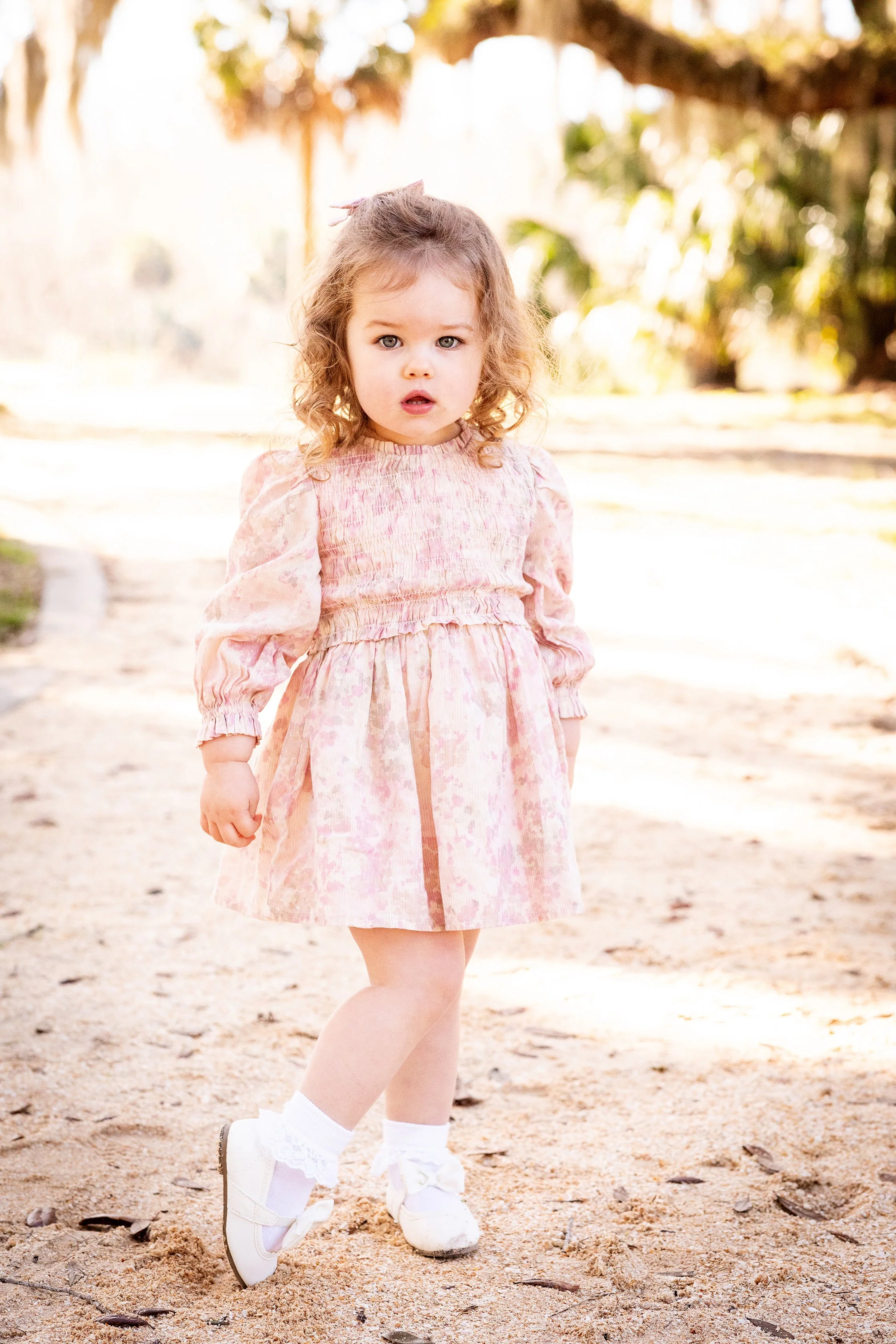 Child portrait of a young girl in a pink dress standing on a sandy path outdoors, in stunning natural light.