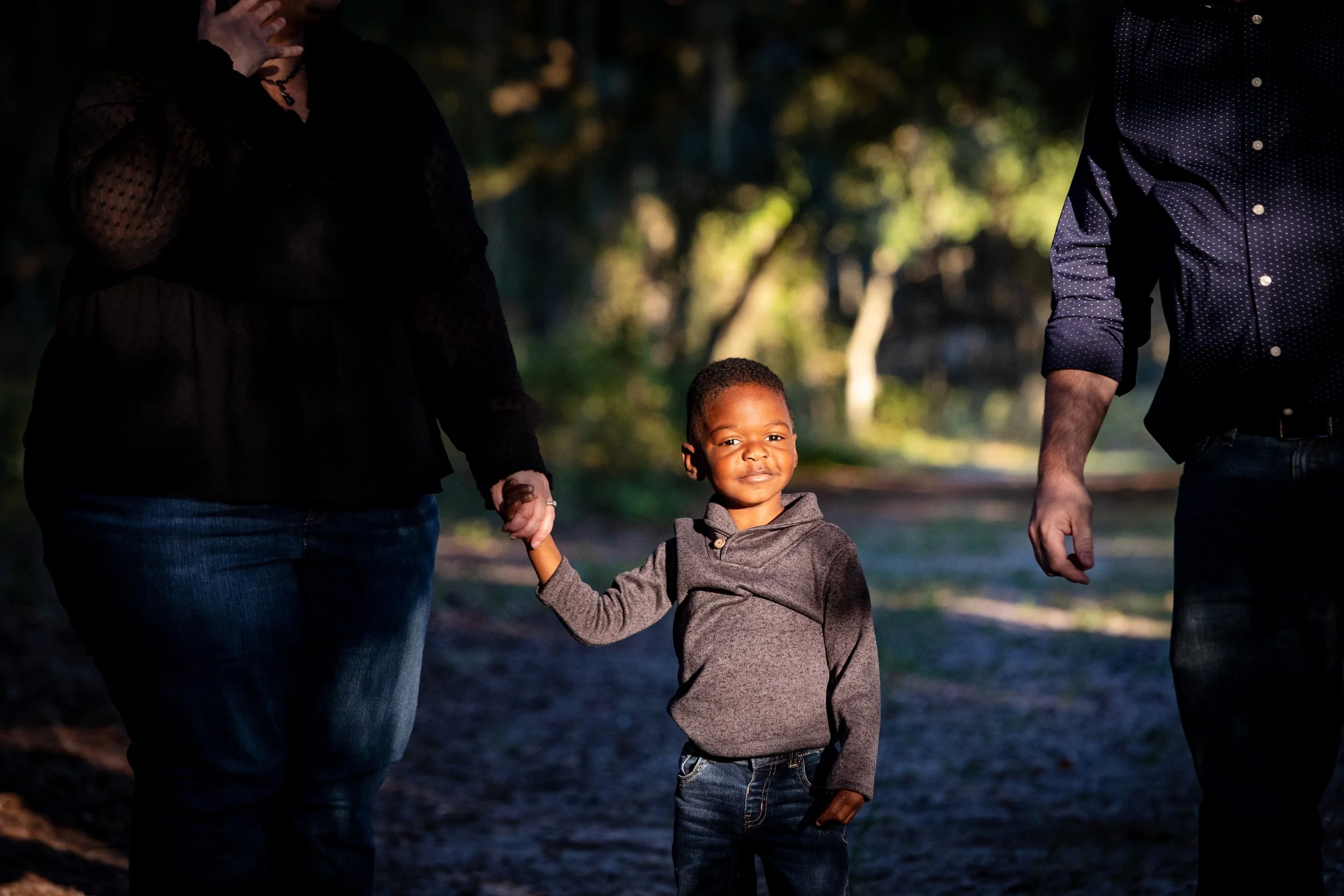 Lifestyle family photography of a young boy holding hands with his parents while walking outdoors under a canopy of Florida trees.