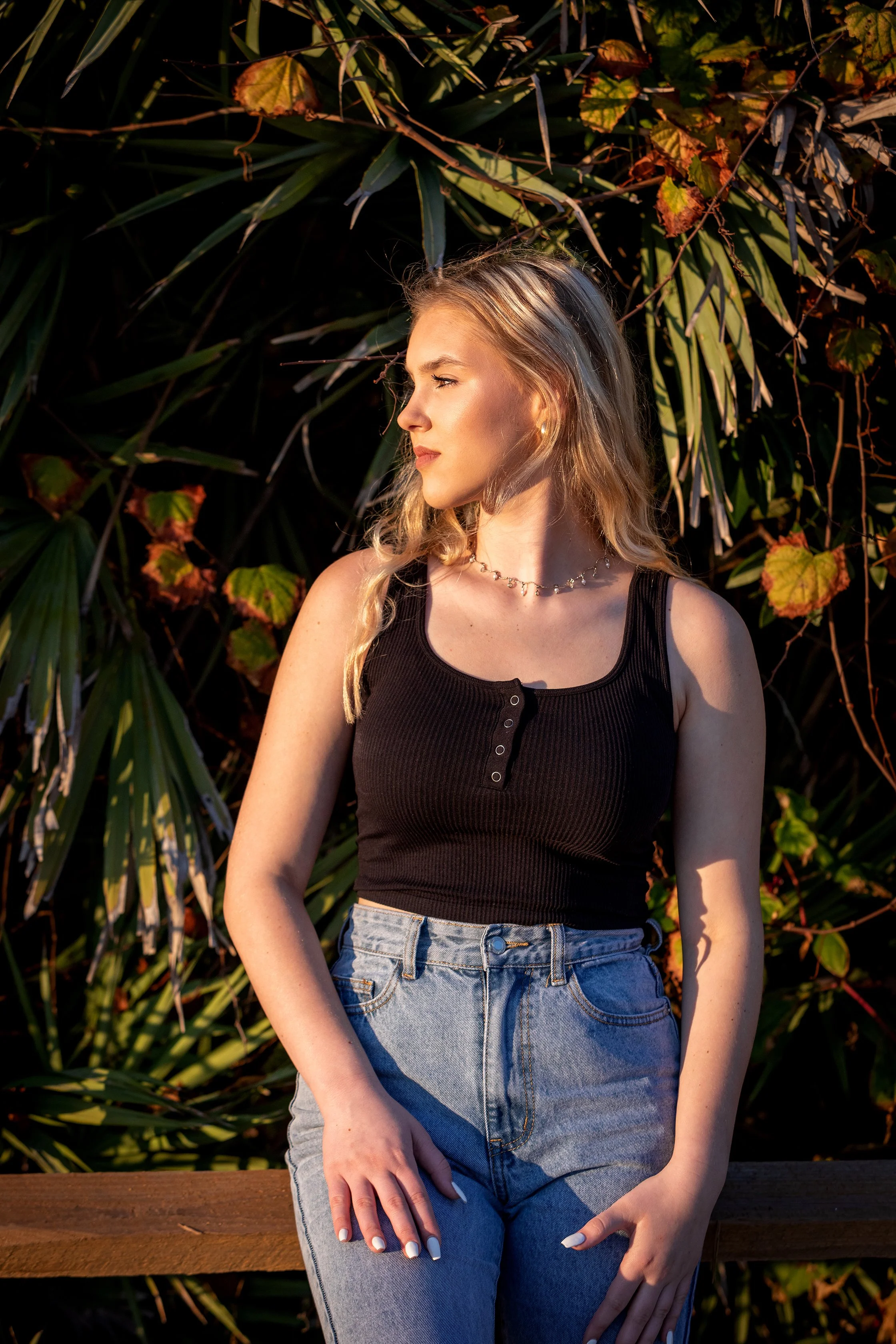 Graduation portrait of a senior wearing a black sleeveless top and high-waisted jeans, standing outdoors near green foliage, illuminated by warm sunlight, photographed by Lindsay Parks.
