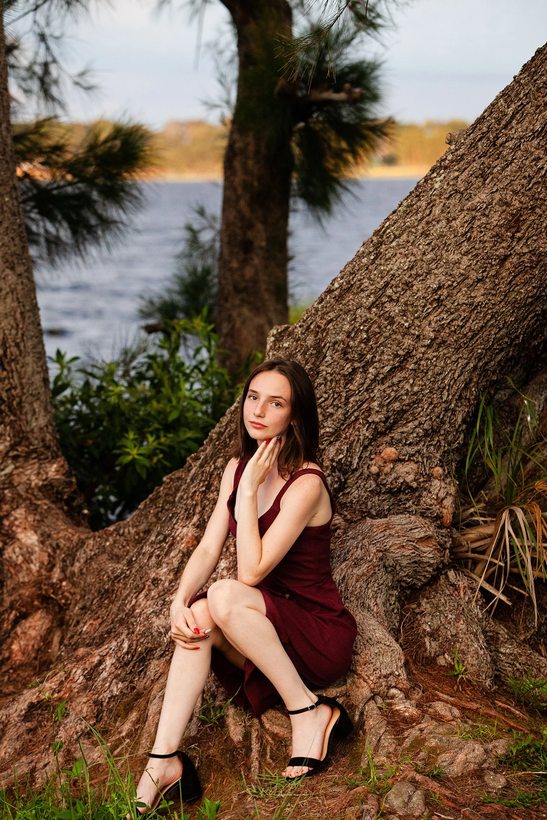 Outdoor portrait of a senior in a burgundy dress seated on tree roots near a lake, photographed by Lindsay Parks Photography