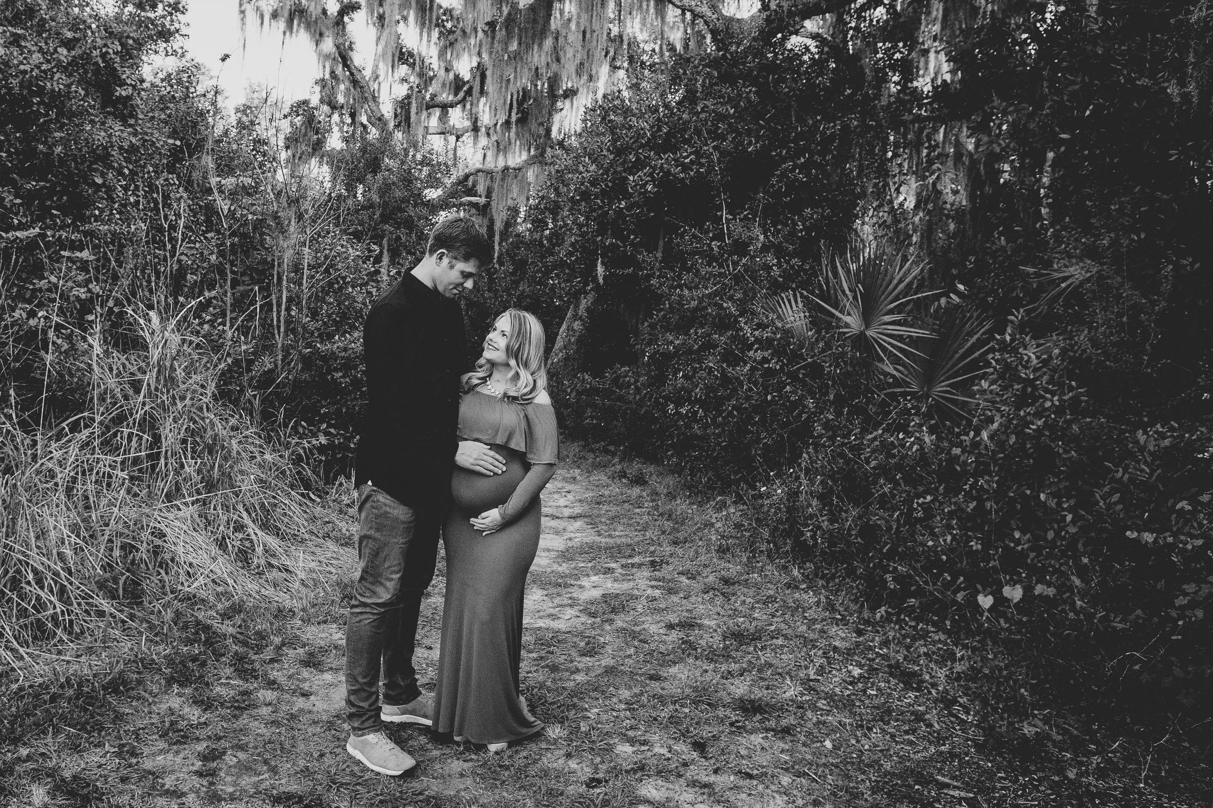 Black and white maternity phophoto of a pregnant woman and a man standing on a dirt path in a wooded area. The woman wears a long dress and is looking up at the man, who is taller and looking down at her, with his hand on her belly.