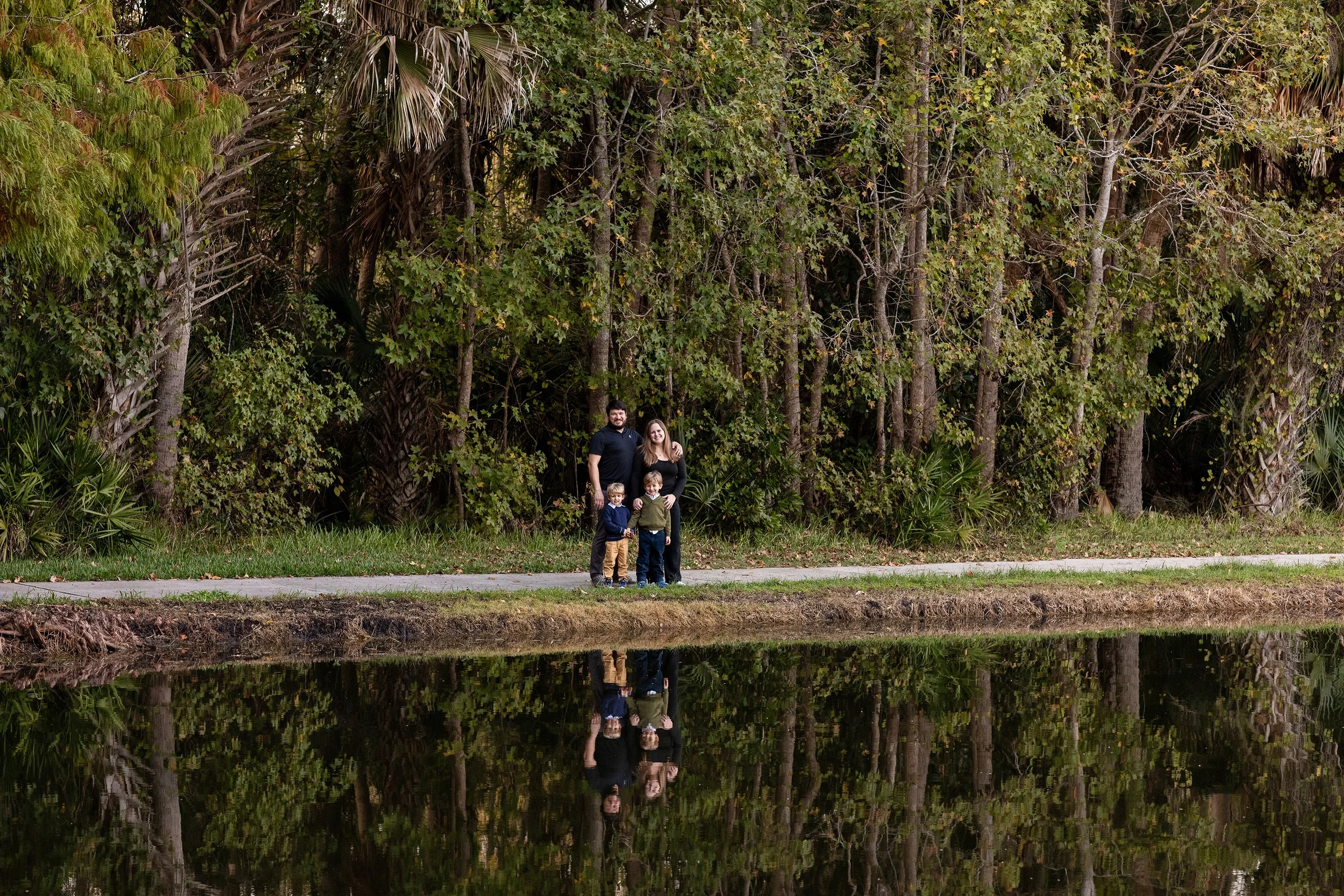 Lifestyle family portrait of a family of four standing beside a pond with stunning reflections in the water with beautiful tall trees in the background.