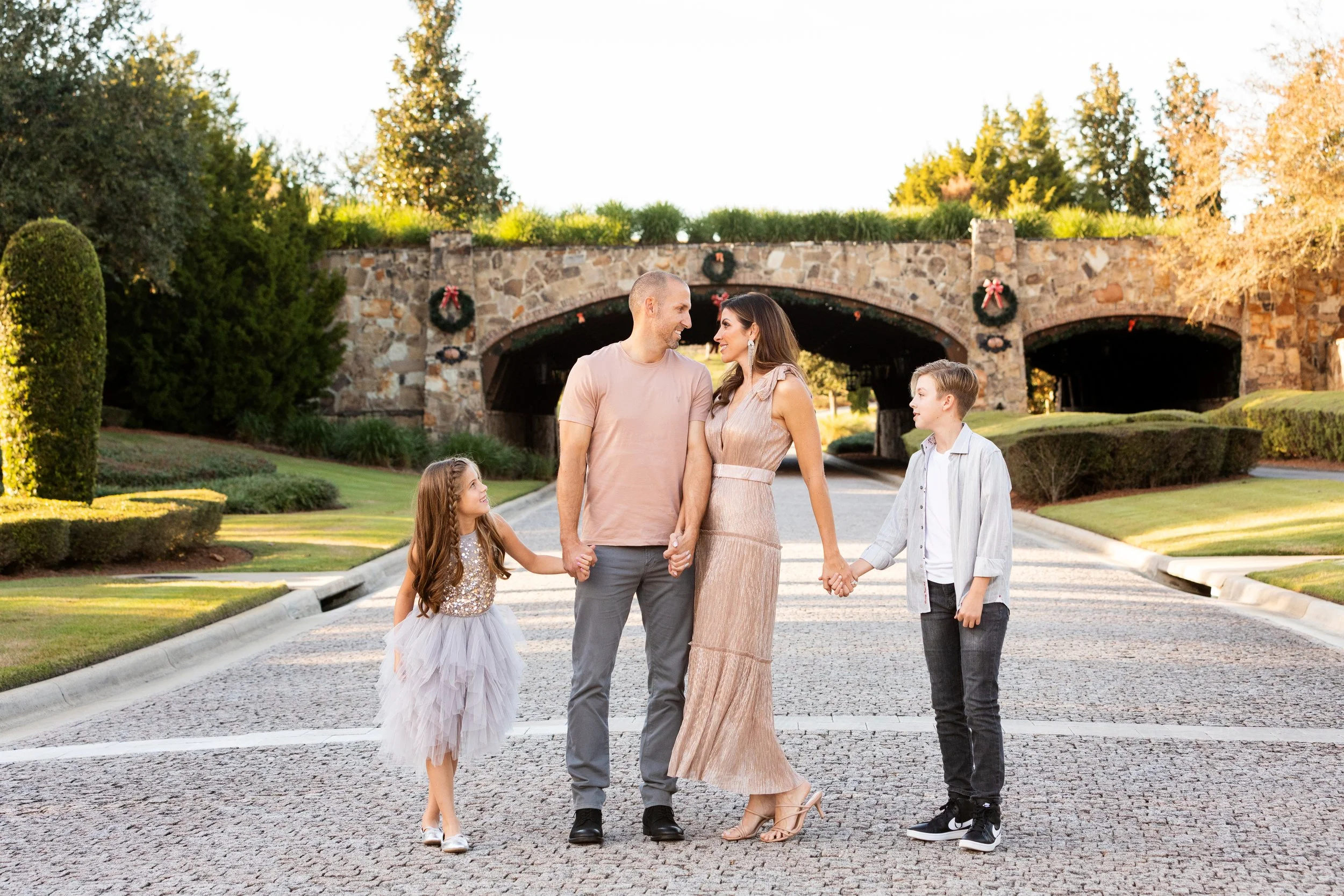 Lifestyle family photography of a family of four holding hands on a cobblestone street, with a stone bridge and Christmas wreath decorations in the background during daytime.