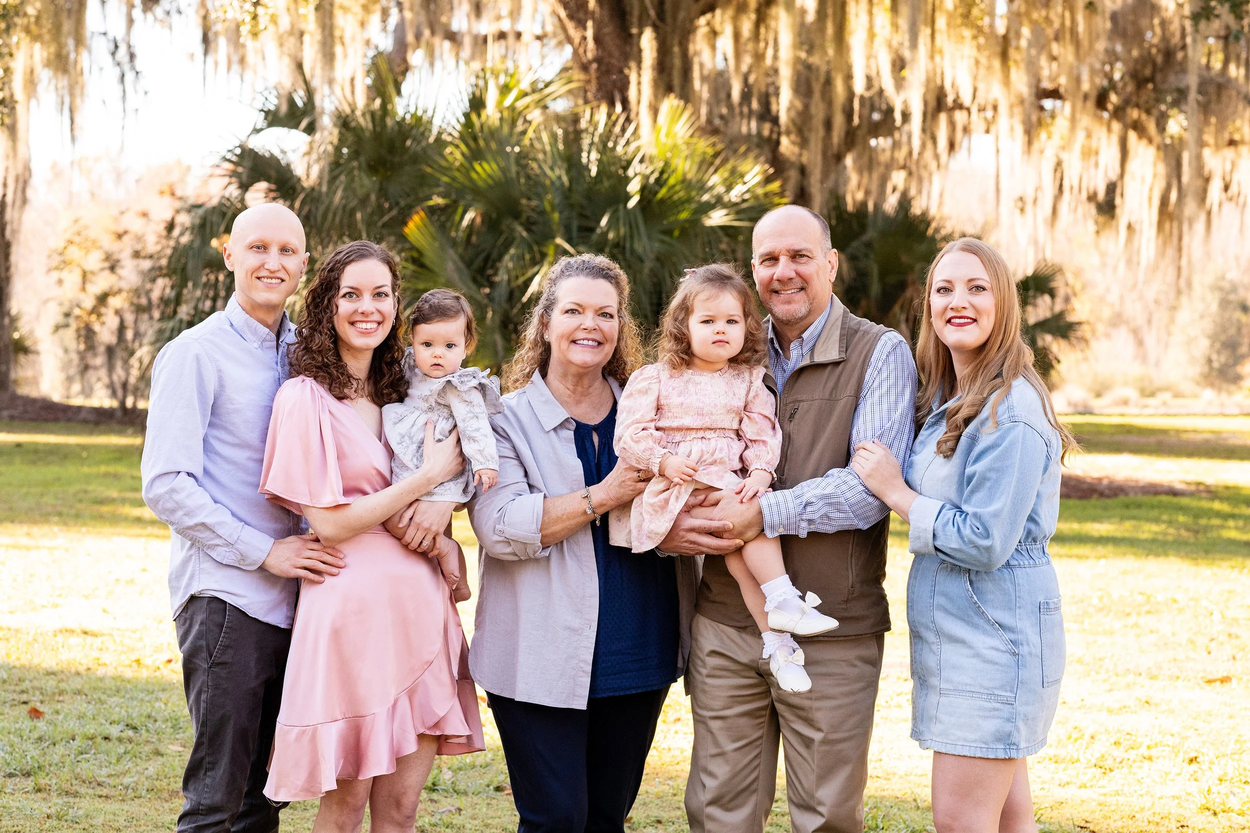 Extended family portrait of seven people gathered outdoors beneath sunlit trees in the beautiful natural light of Florida golden hour.