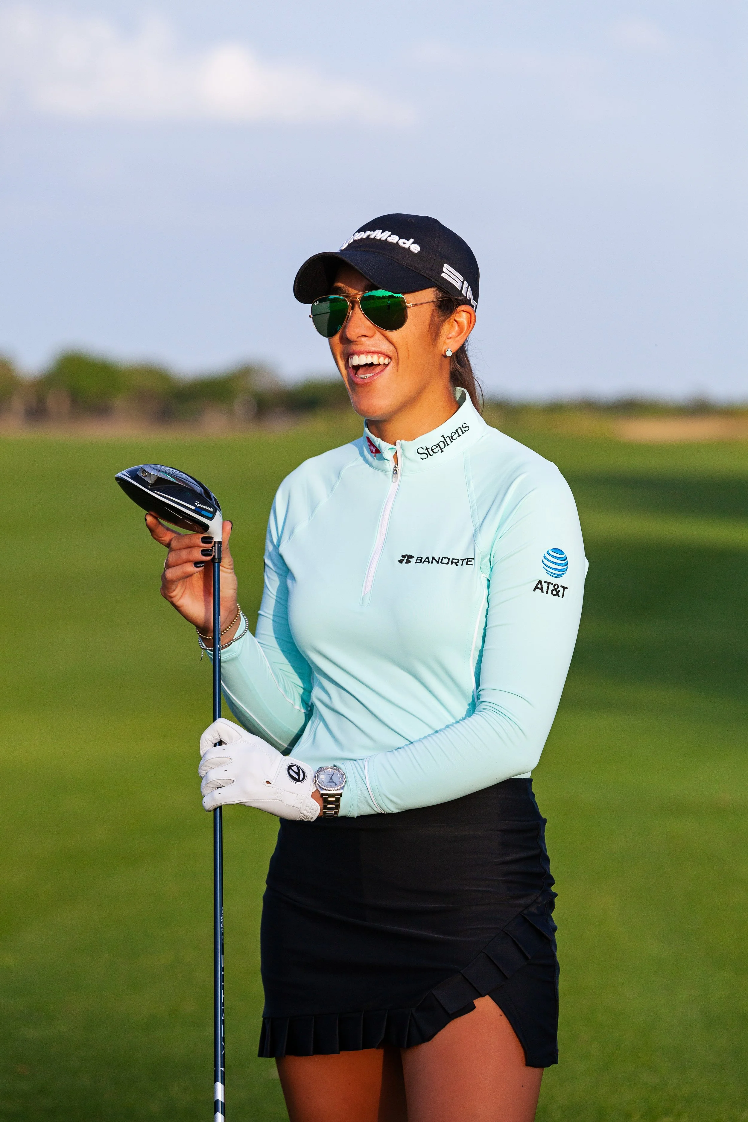 Lifestyle golf portrait photography of a woman wearing a cap and sunglasses, holding a club, and smiling on a golf course.