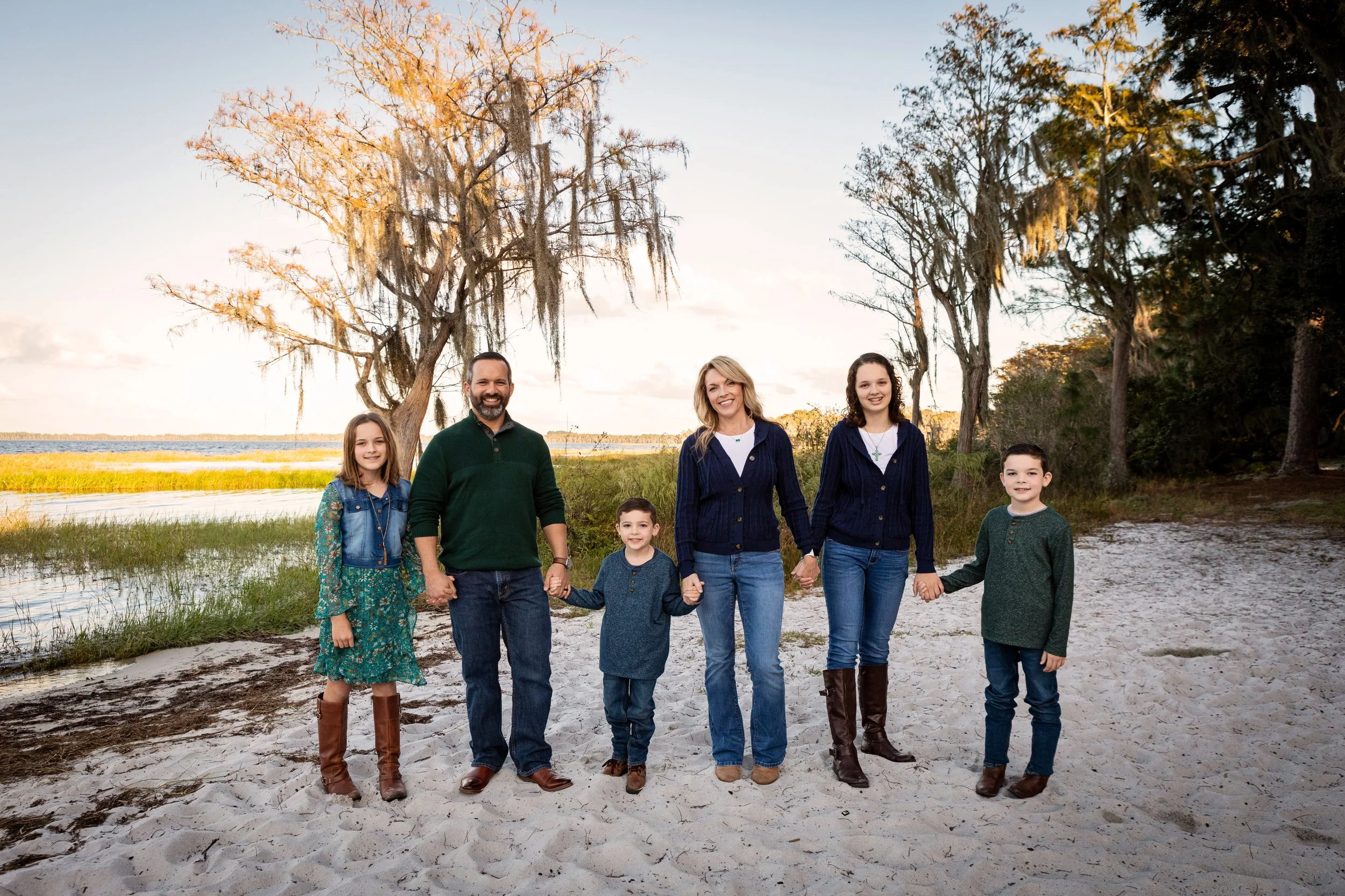 Family photography of a family of seven standing hand-in-hand on a sandy beach near a body of water, with trees and calm water in the background during sunset.