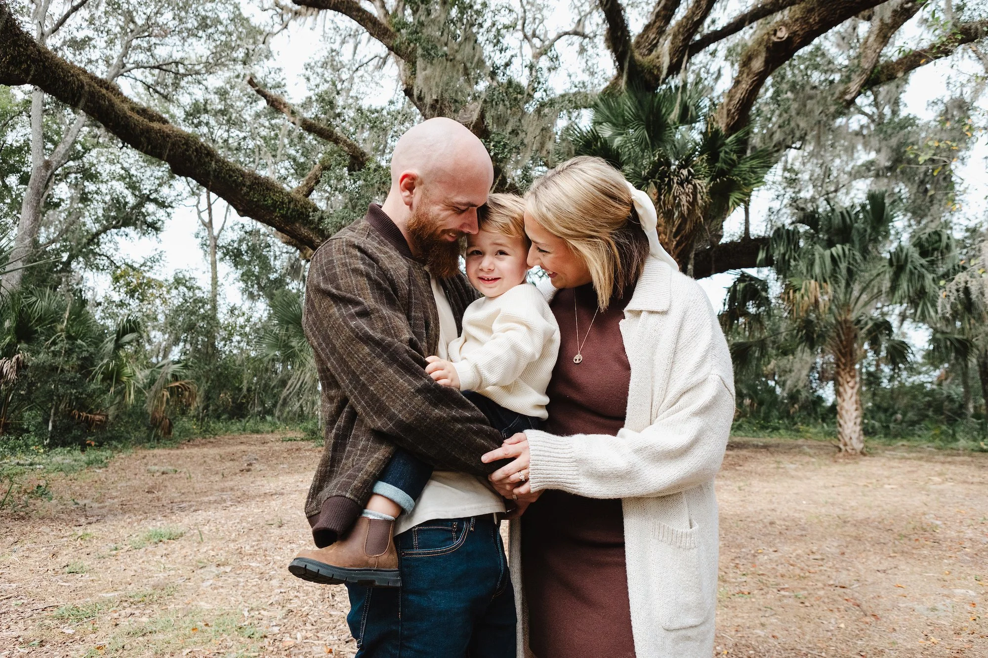 Outdoor family lifestyle photography of parents holding their young son beneath a large tree, photographed by Lindsay Parks Photography