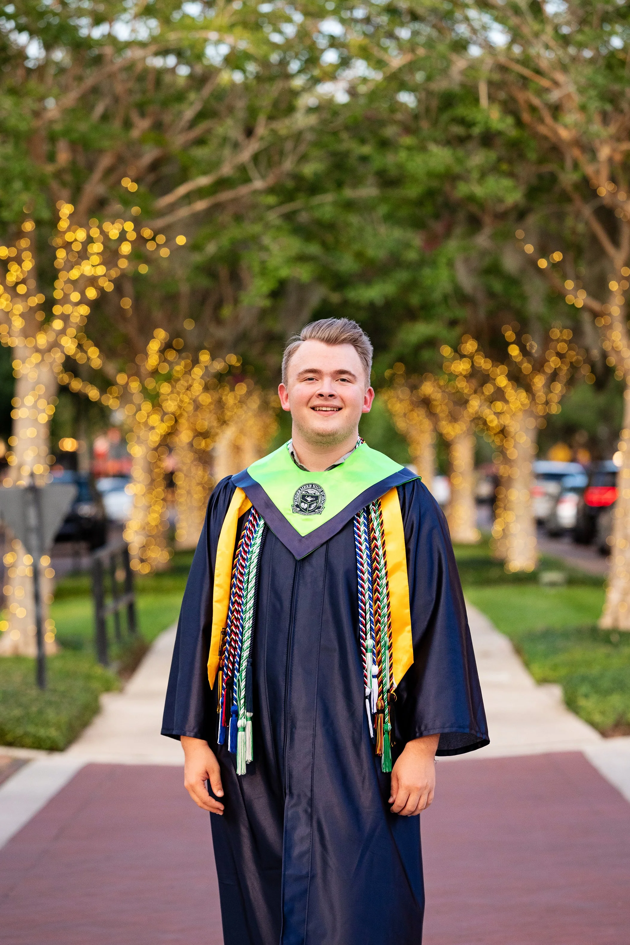 Senior graduation portrait of a young man in cap and gown standing beneath string lights at sunset, photographed by Lindsay Parks Photography