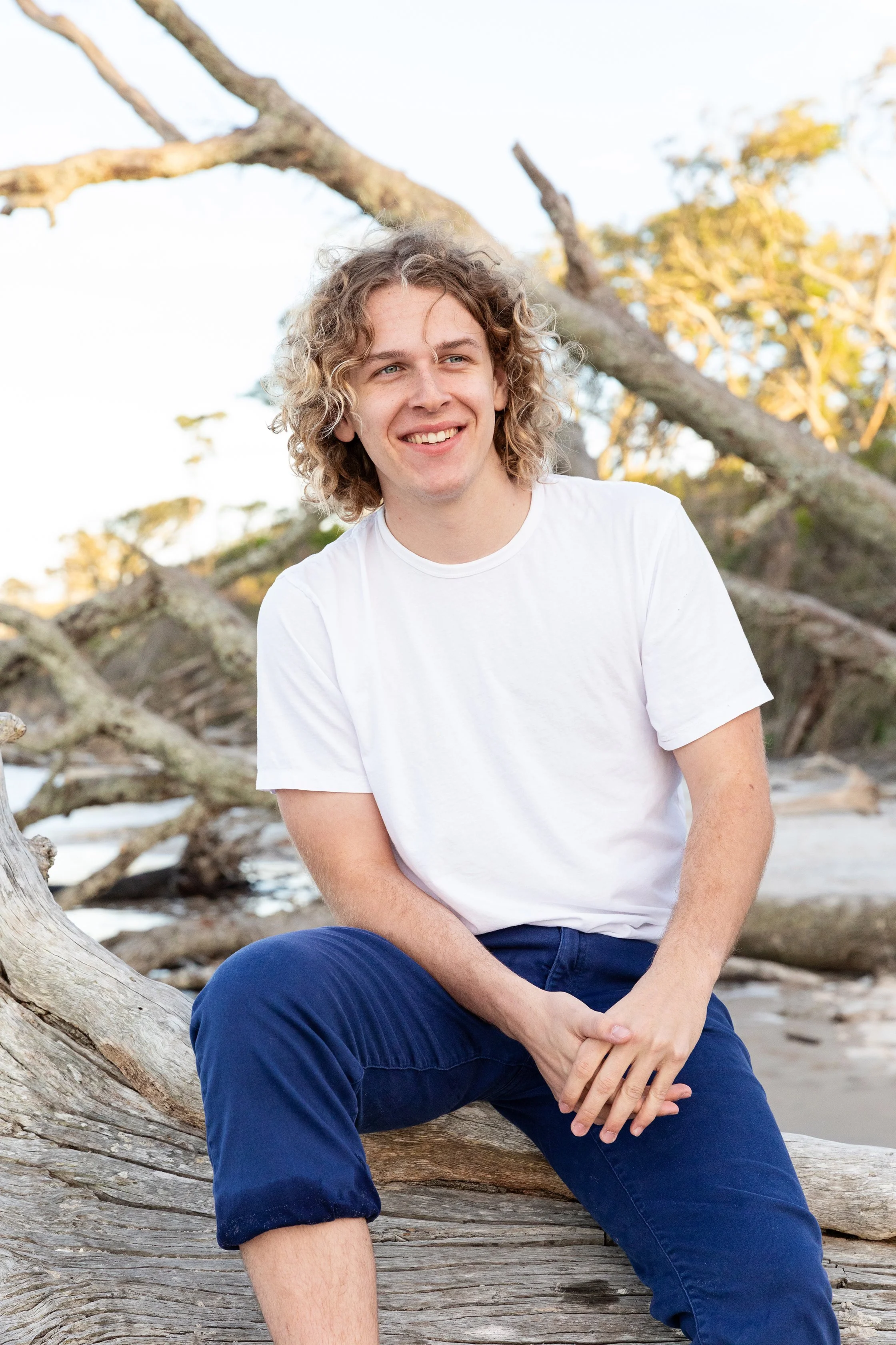 Lifestyle portrait of a young man seated on a fallen tree trunk on the beach at sunset, photographed by Lindsay Parks Photography