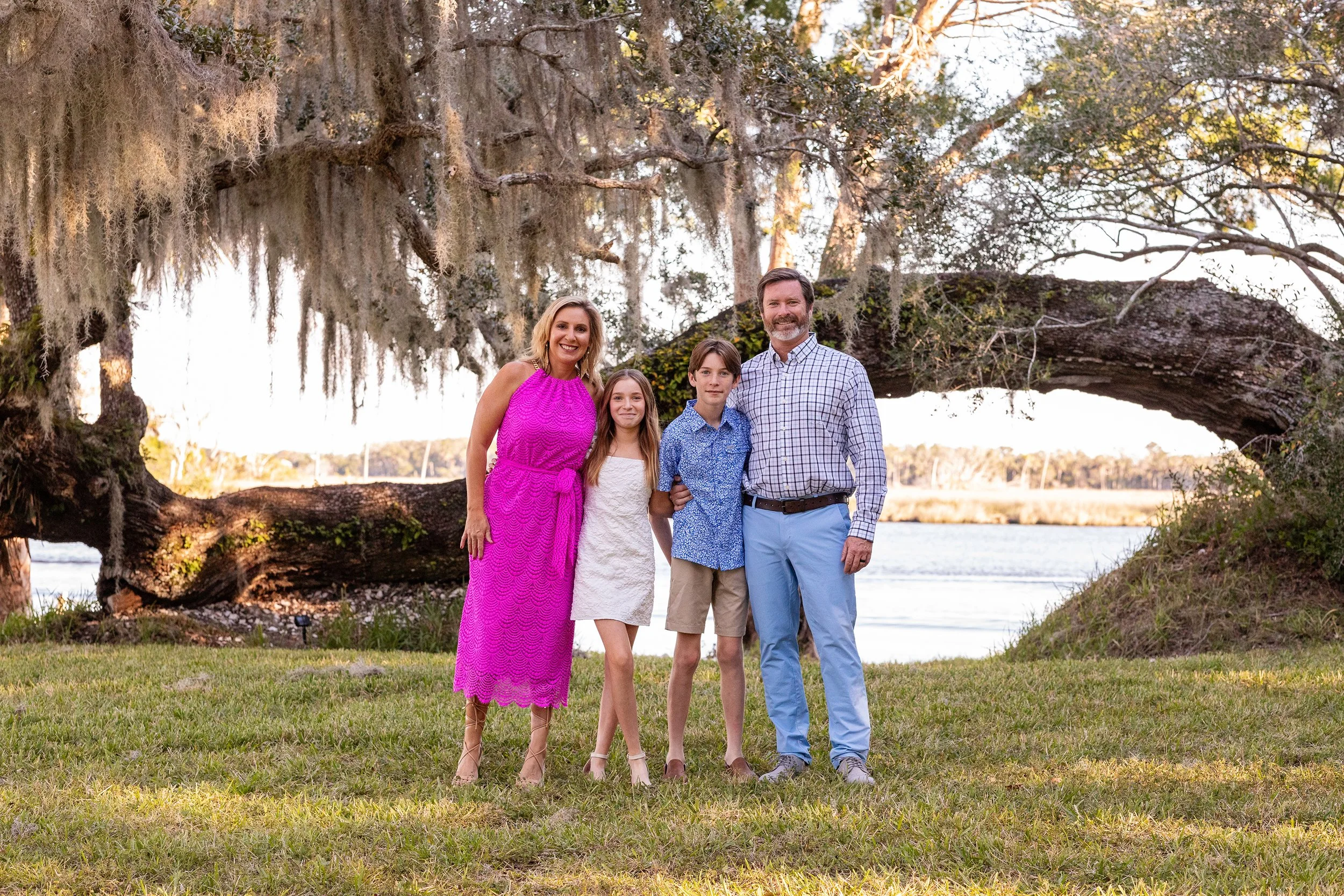 Outdoor family portrait of parents and children standing near a waterway with hanging moss, with a large tree, during daytime. 