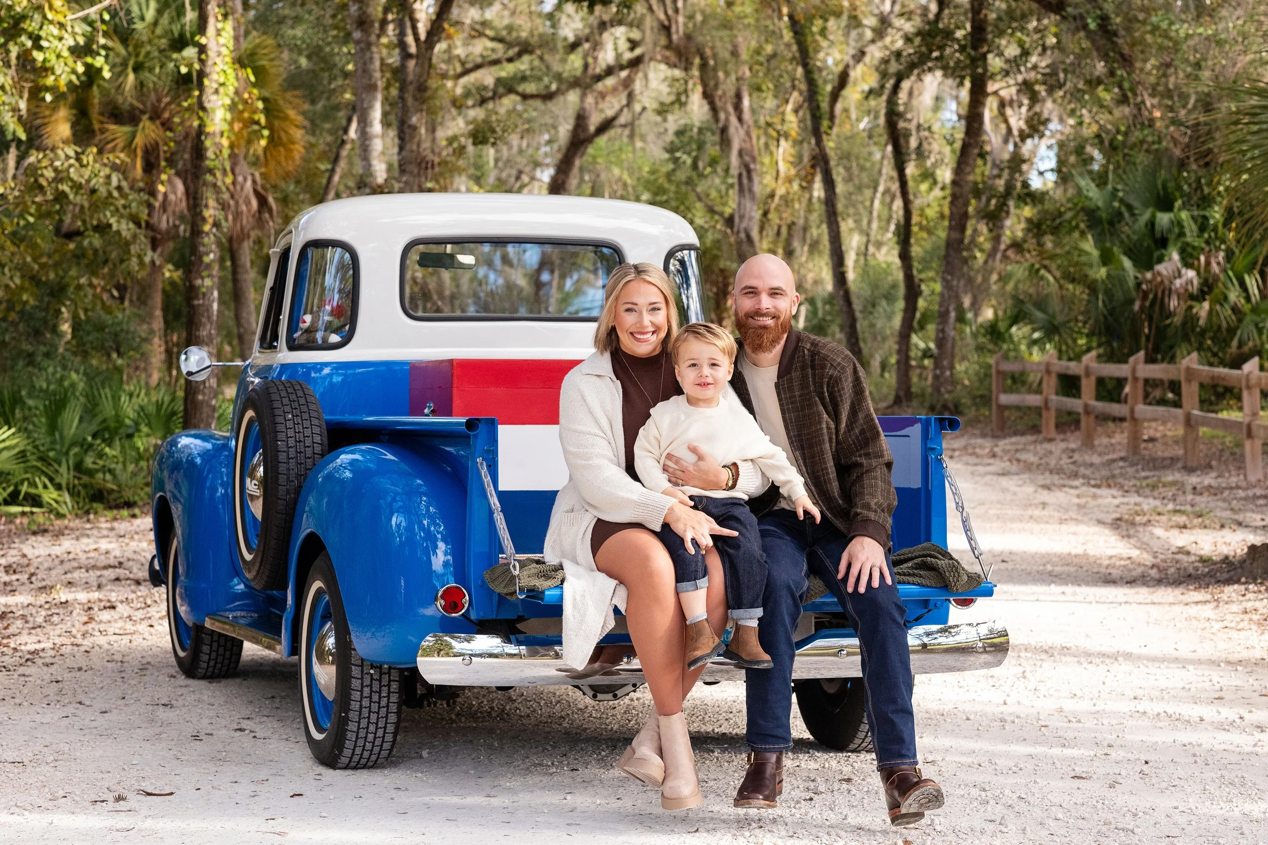 Lifestyle family portrait of a family of three sitting on a vintage pickup truck outdoors in a wooded area, smiling at the camera.