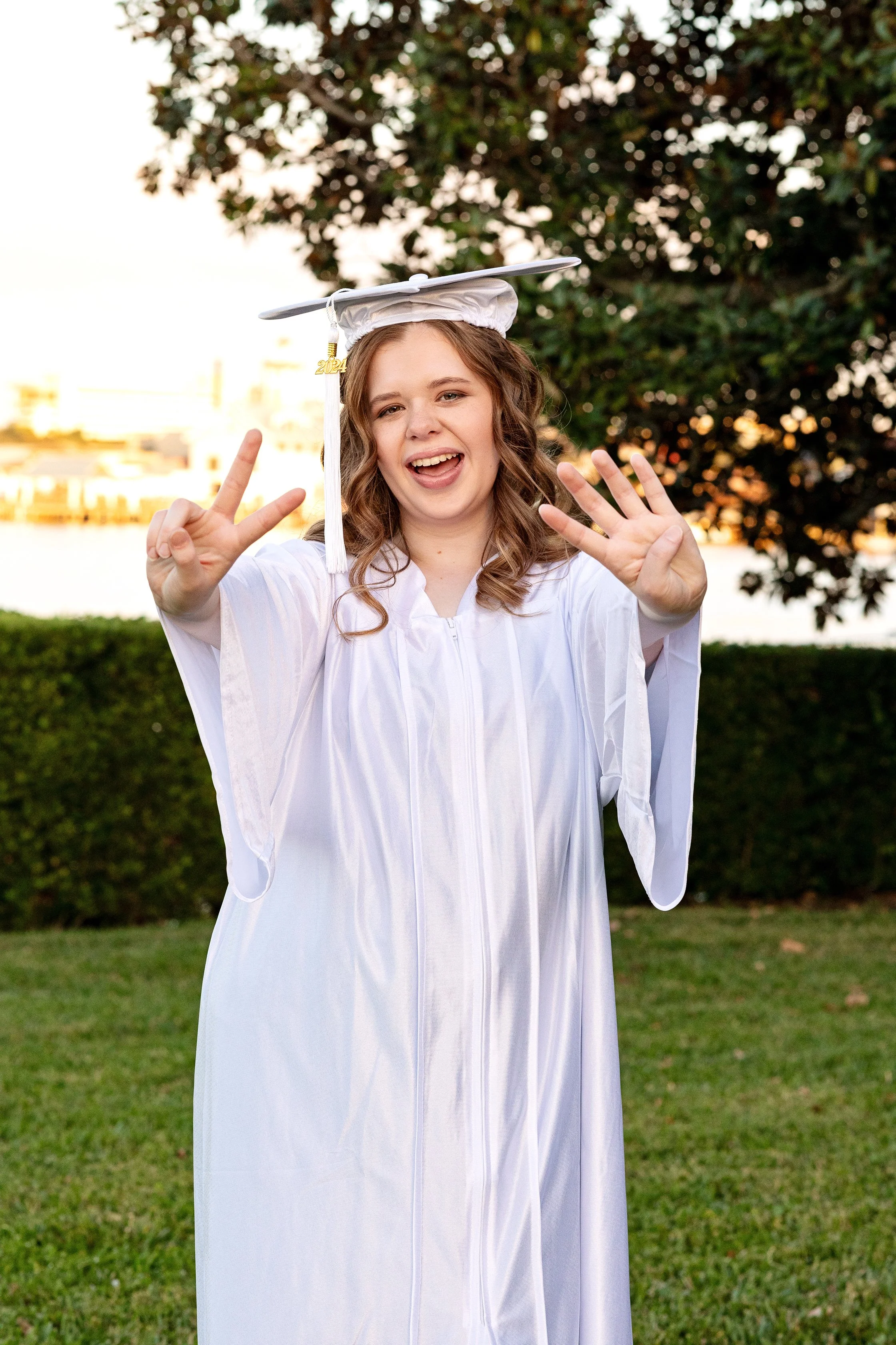 Graduation photography of a young woman celebrating in a white cap and gown near water at sunset