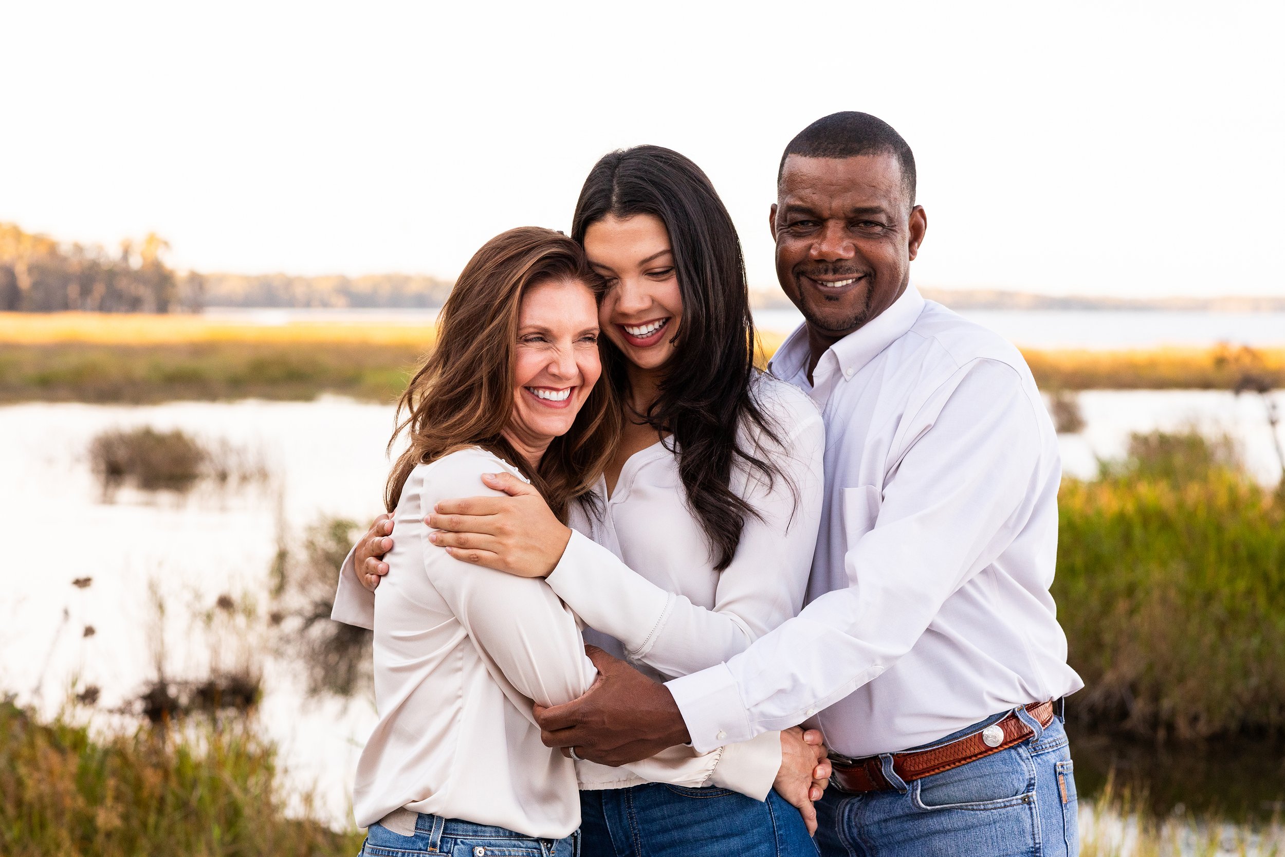 Family lifestyle photography of three people hugging near water and tall grasses at sunset, photographed by Lindsay Parks Photography