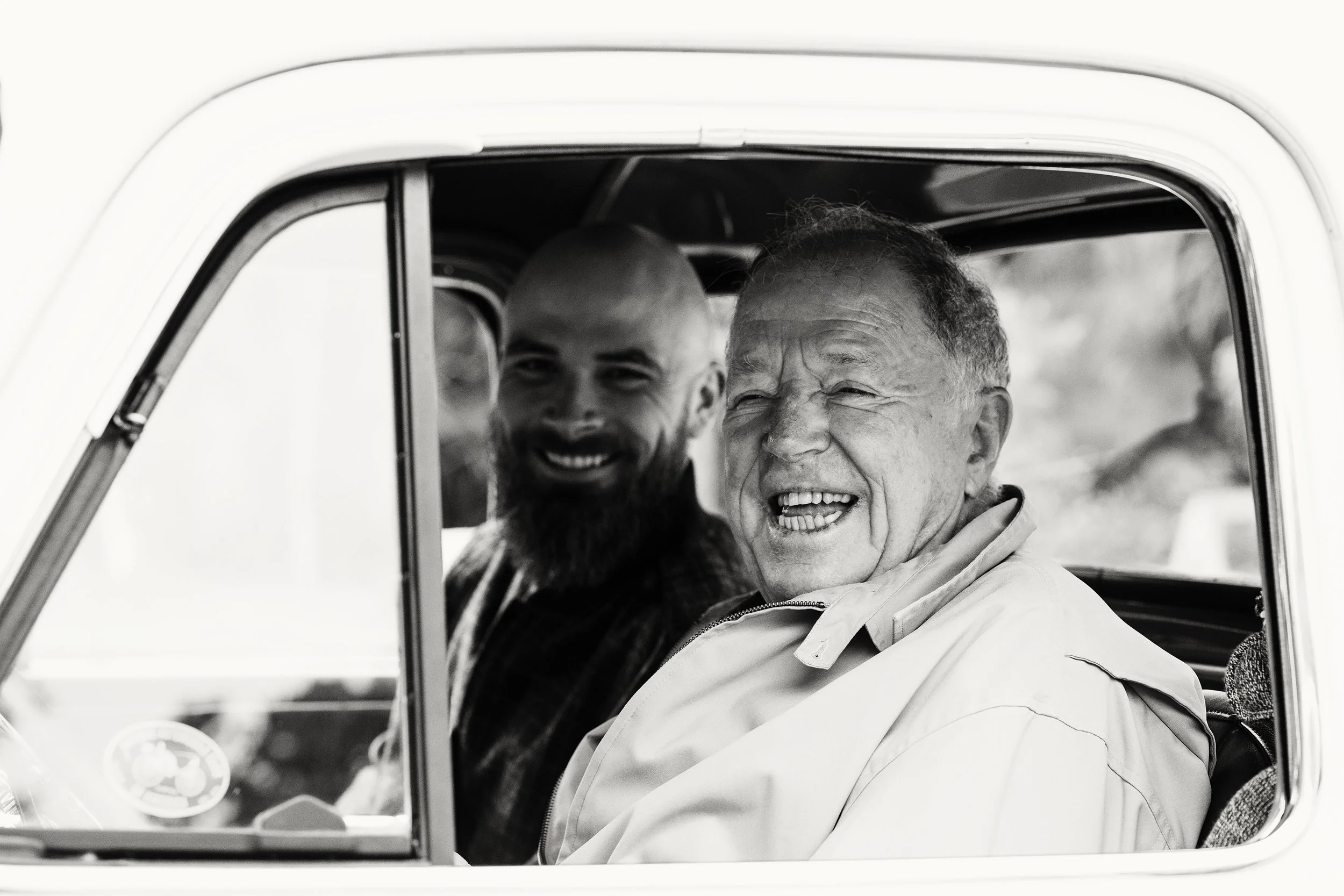 Lifestyle portrait of two men smiling together while seated inside a truck. One elderly with a light jacket, and a younger man with a beard and bald head, both appearing happy and enjoying the moment.
