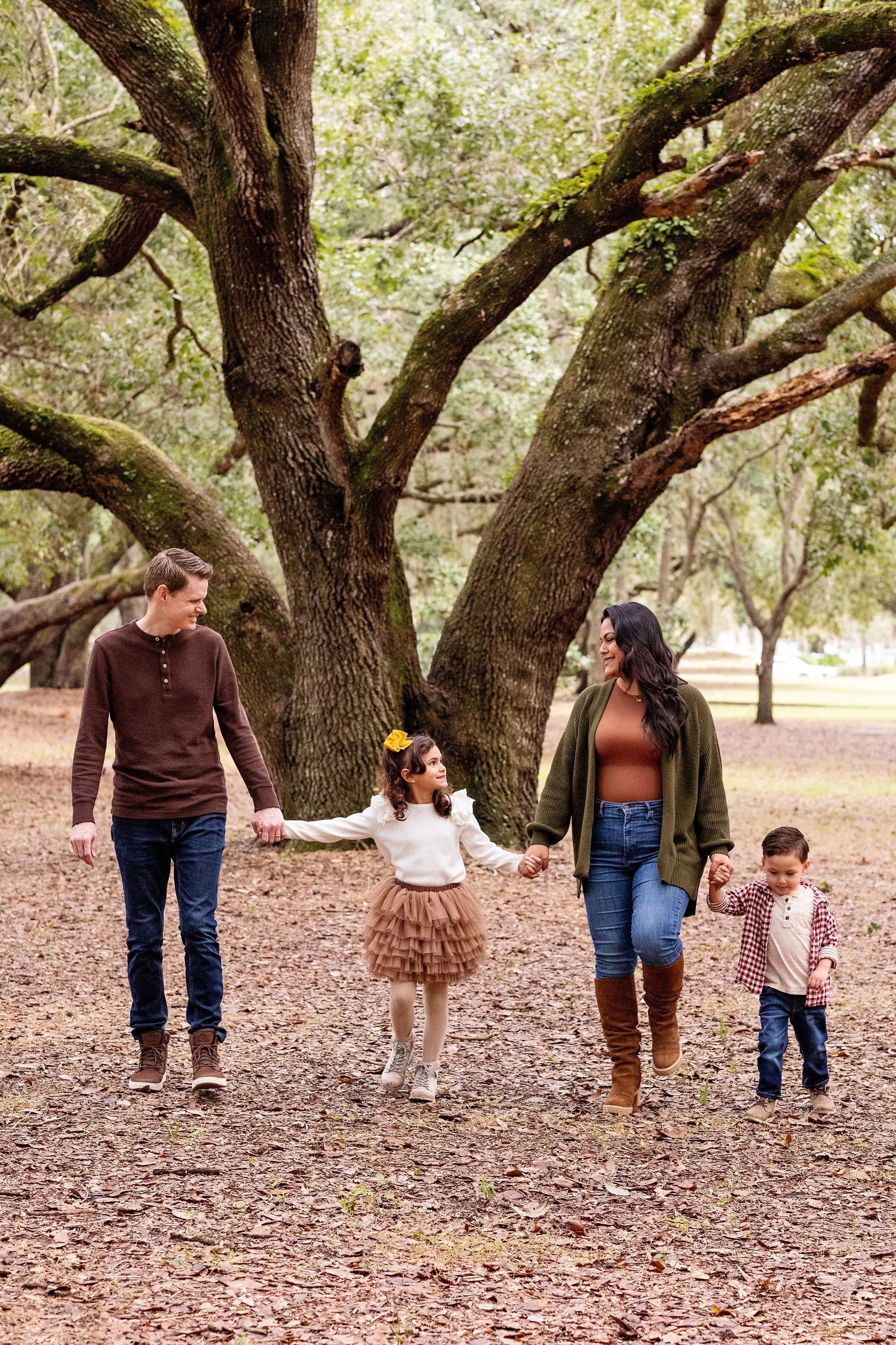 Fall family photography of a family of four walking hand in hand beneath large trees and surrounded by scattered leaves on the ground.