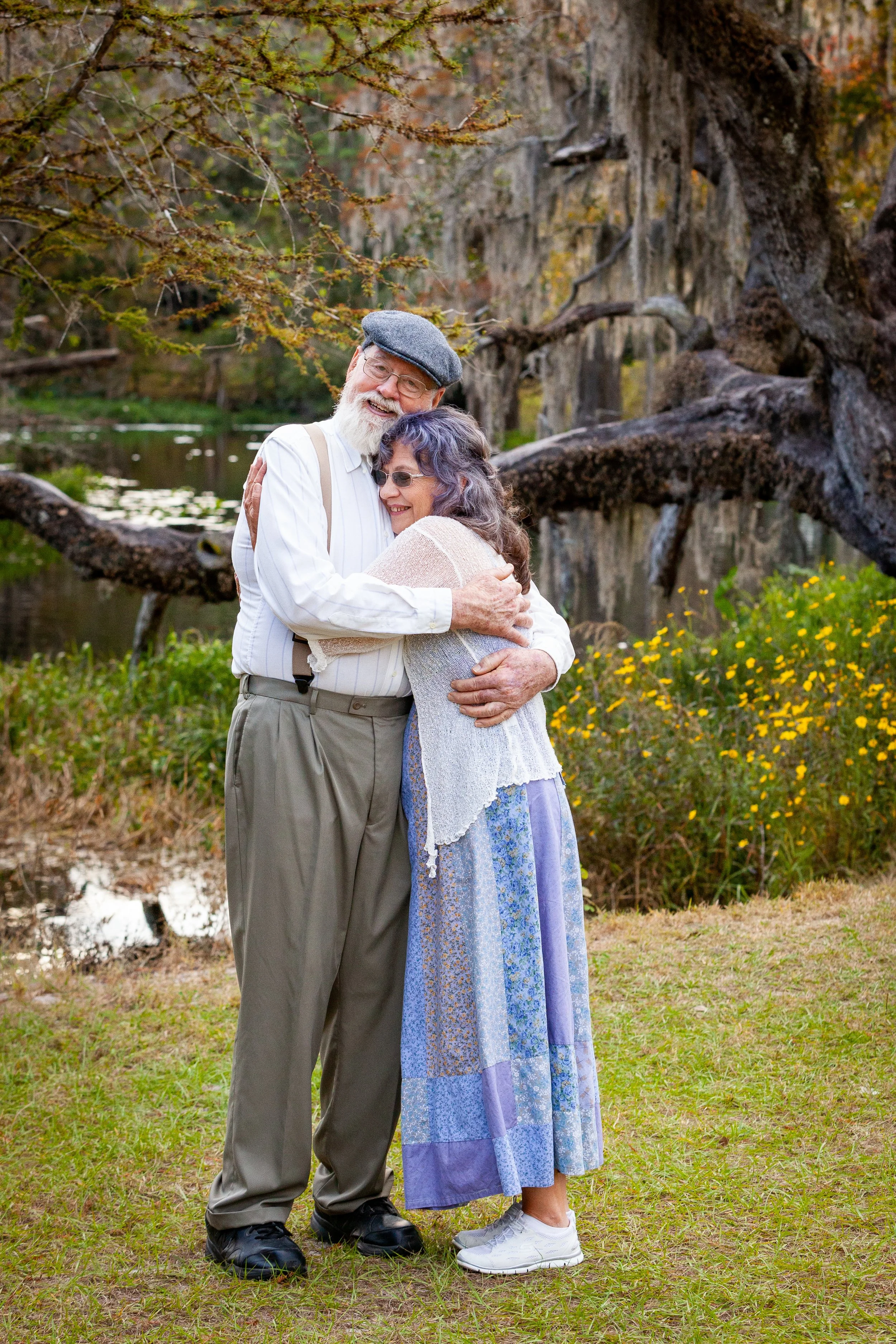Lifestyle portrait of an elderly couple sharing a warm hug beside a pond in autumn, surrounded by trees with green foliage and yellow flowers.