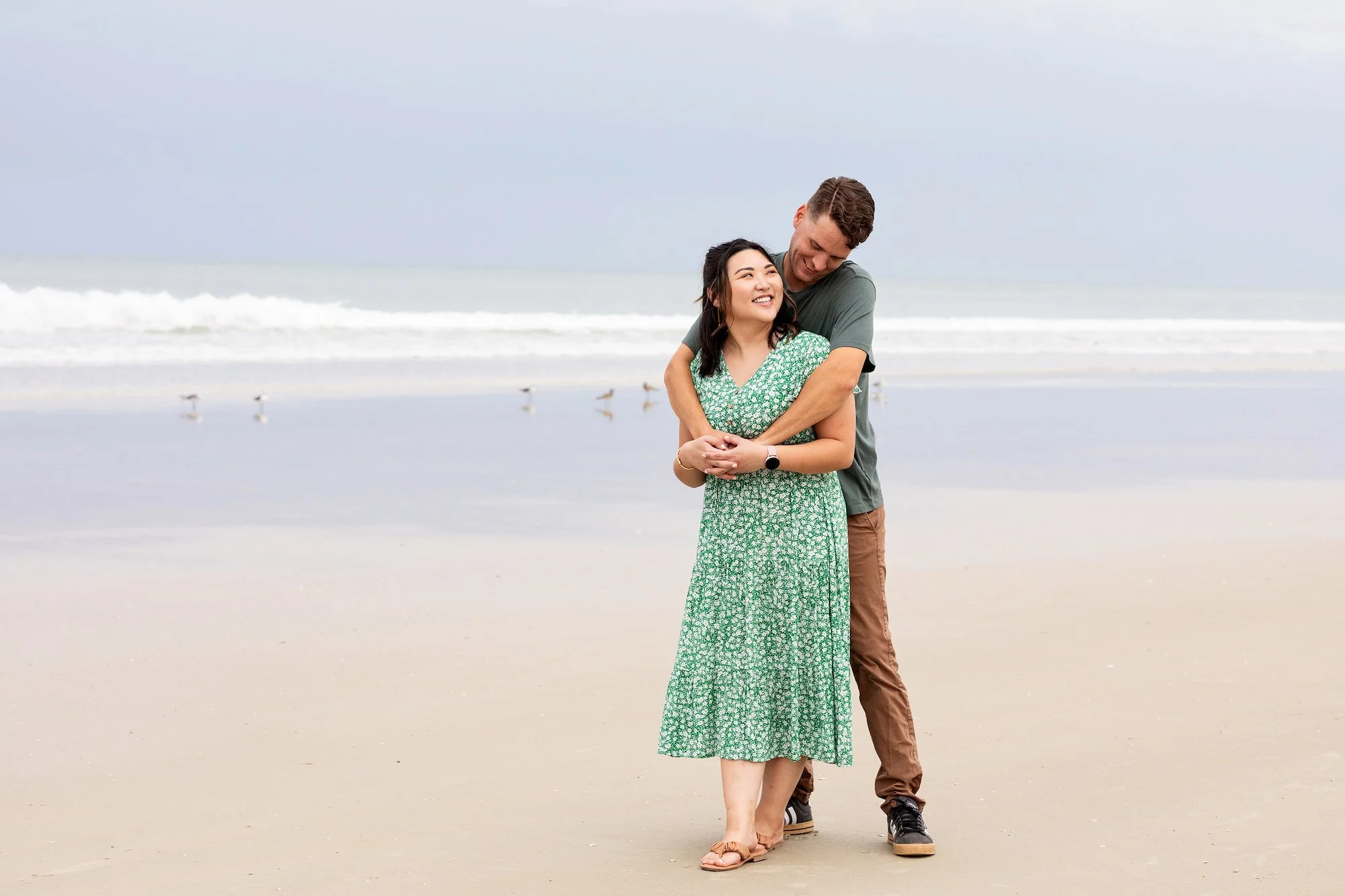 Portrait photography beach engagement photo of a couple hugging and smiling with ocean waves and seagulls, photographed by Lindsay Parks Photography on a Florida beach.
