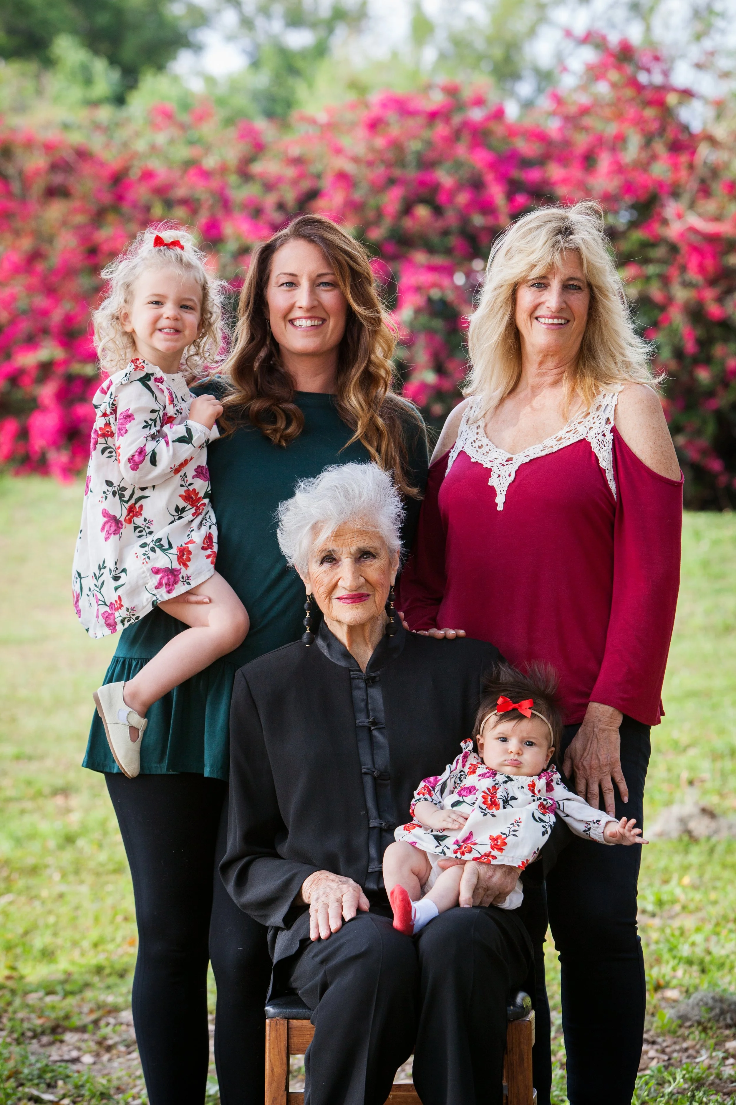 A multigenerational family portrait photo featuring five women and two young girls outdoors with pink flowering bushes in the background. The elderly woman is seated holding a baby, and two women are standing behind them.