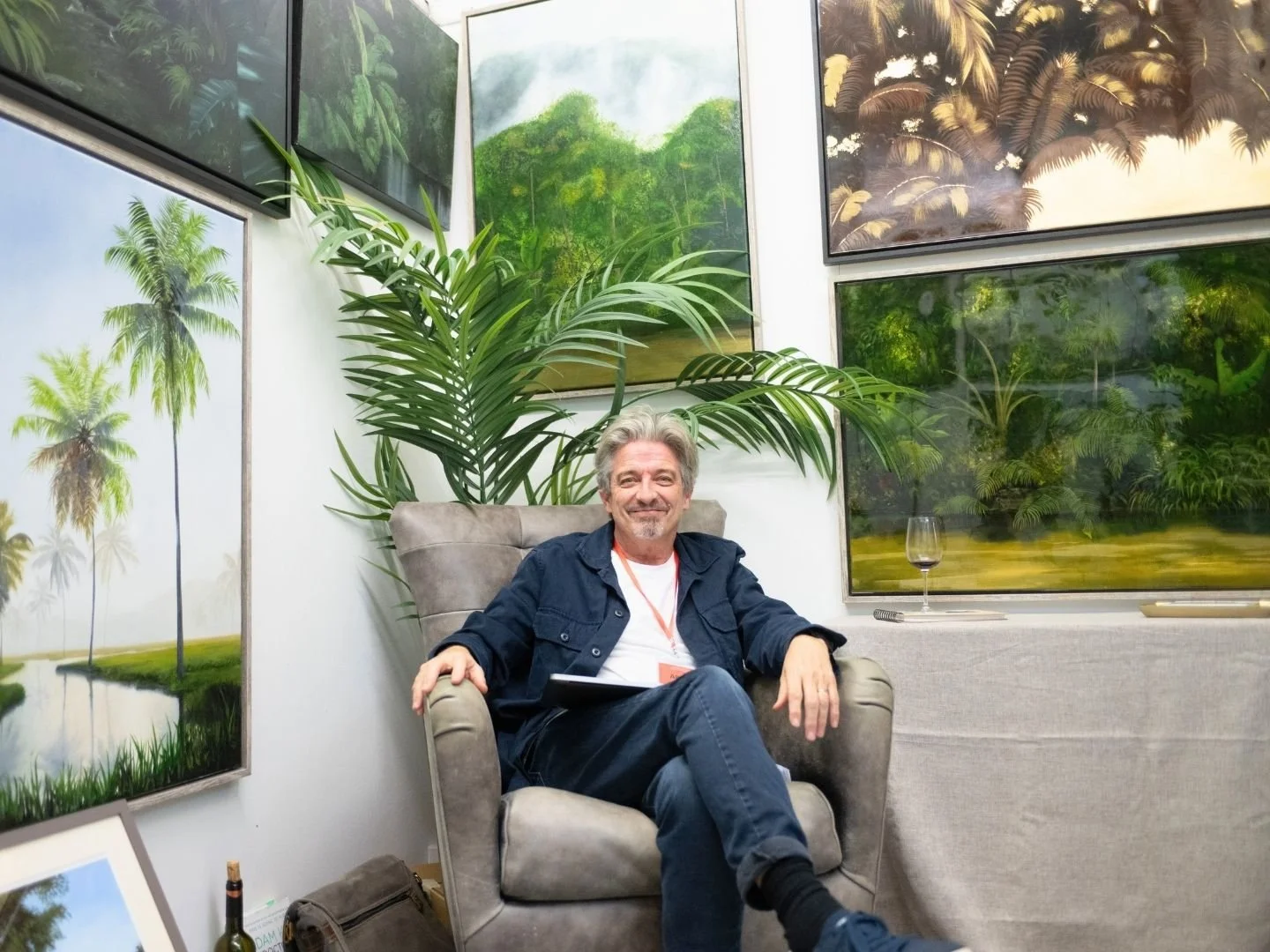 A man with gray hair and a gray beard sitting in a beige armchair, smiling. He is wearing a dark jacket, white shirt, and dark pants. Behind him is a large green plant, and he is surrounded by framed landscape paintings featuring tropical scenes with palm trees and lush greenery on the walls.