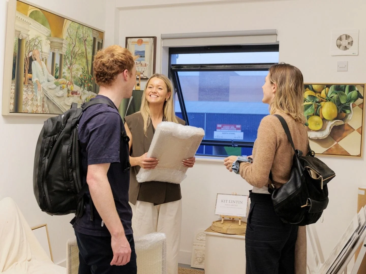 Three people standing and talking inside an art gallery, with paintings hanging on the walls and a window in the background. One woman is holding a rolled-up piece of bubble wrap.