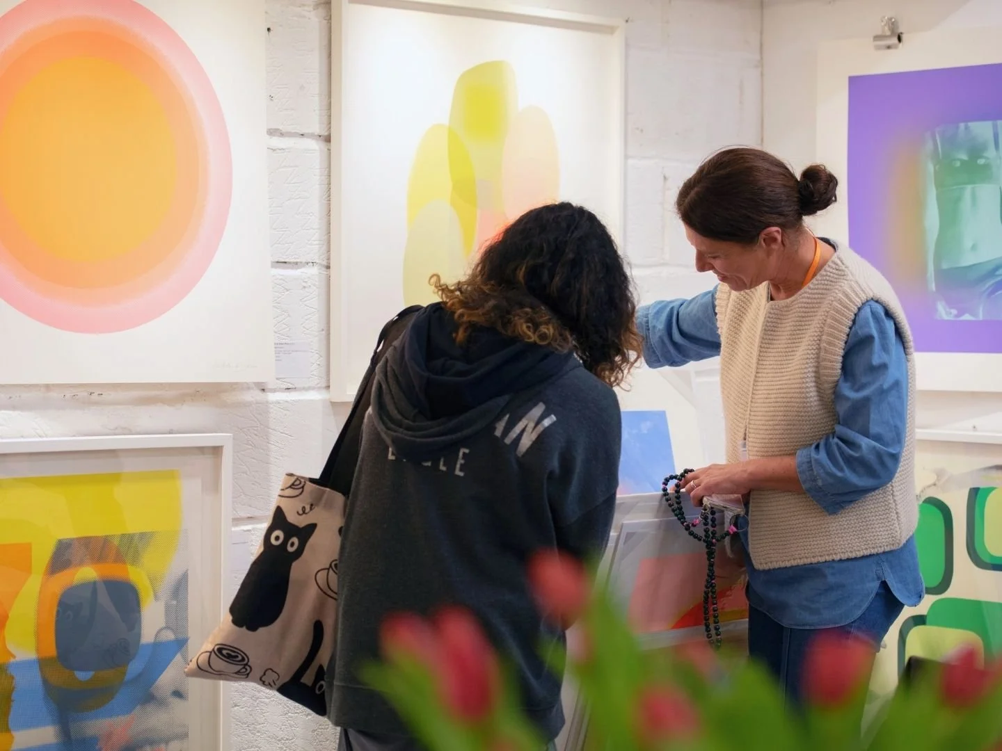 A studio visitor and a printmaker artist in her art studio looking at colourful abstract prints on the wall. One woman is holding a beaded necklace or bracelet, and the other has a black tote bag with a black cat illustration.