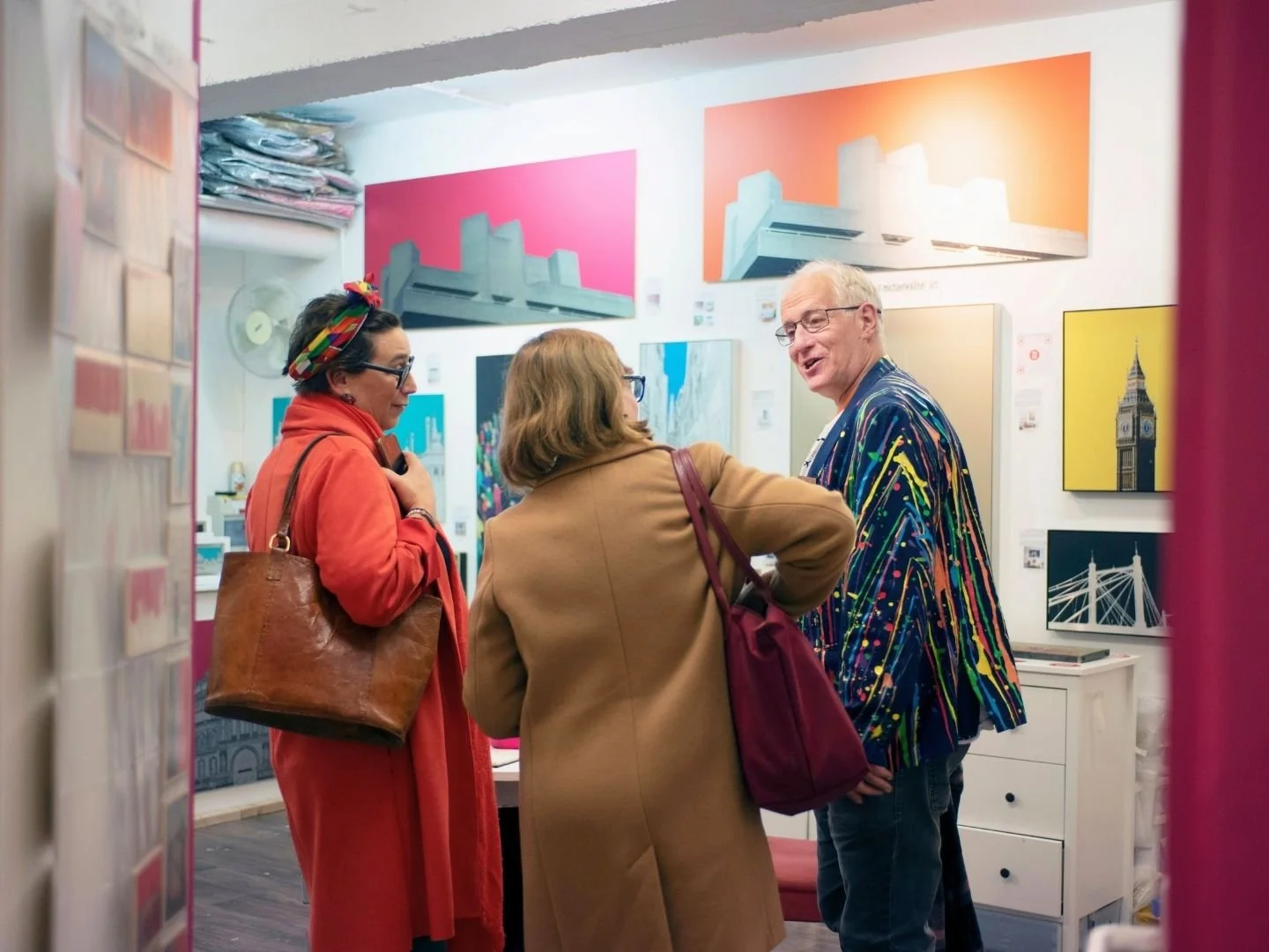 Three women and one man standing and talking in an art studio, surrounded by colorful abstract paintings and photographs of landmarks.