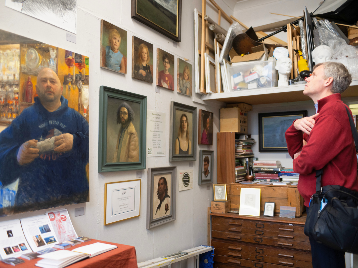 Two men are viewing artwork inside an art studio. One man on the left is reflected in a mirror, and the other on the right stands with arms crossed looking at framed portraits and paintings on the wall.