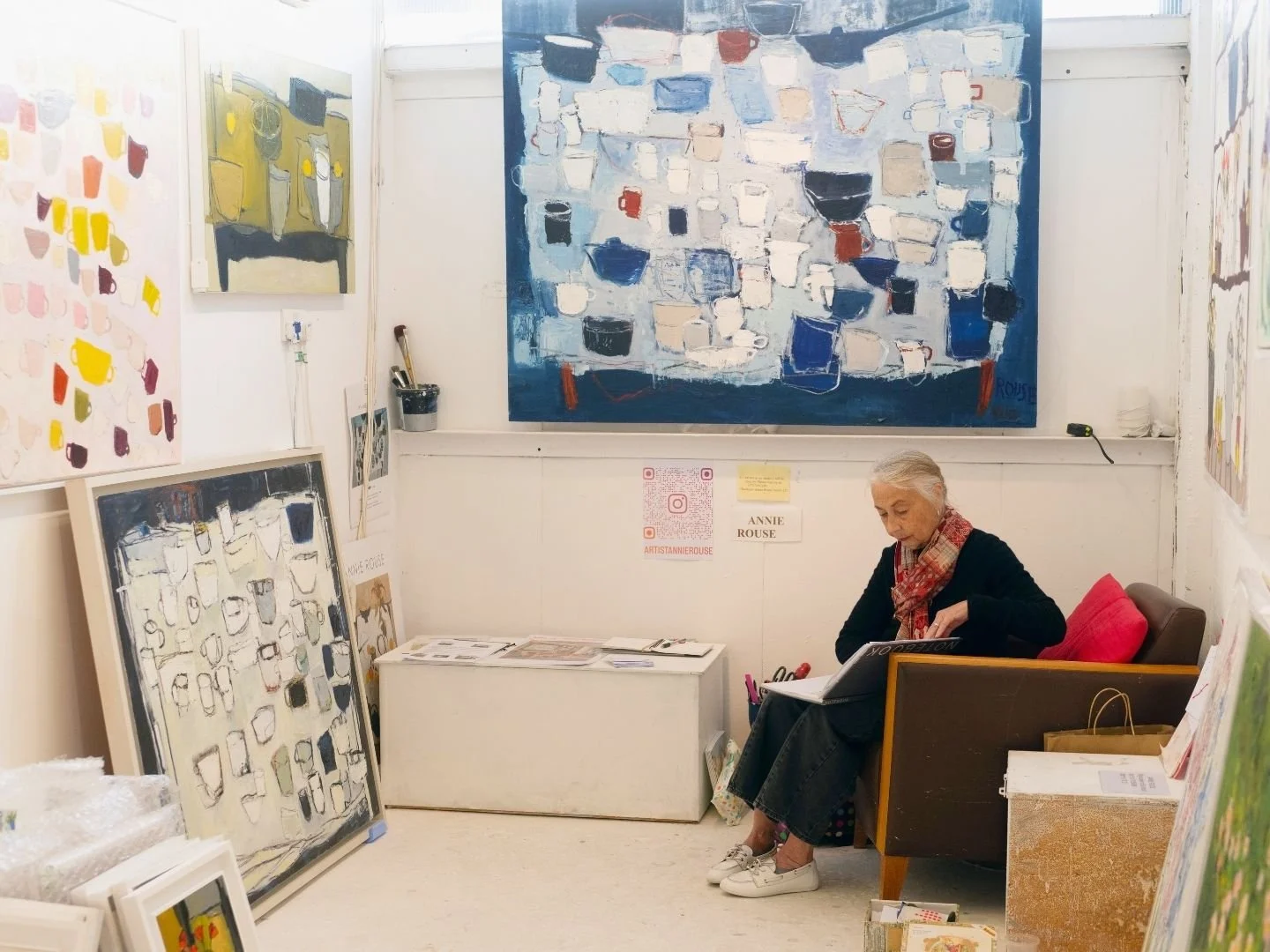 An elderly woman sits on a brown chair in an art gallery or studio, surrounded by colorful abstract paintings on the walls and leaning against the floor. She is wearing a black sweater, a patterned scarf, and white sneakers, reading a book. The room has white walls and small signs and QR codes on the wall behind her.