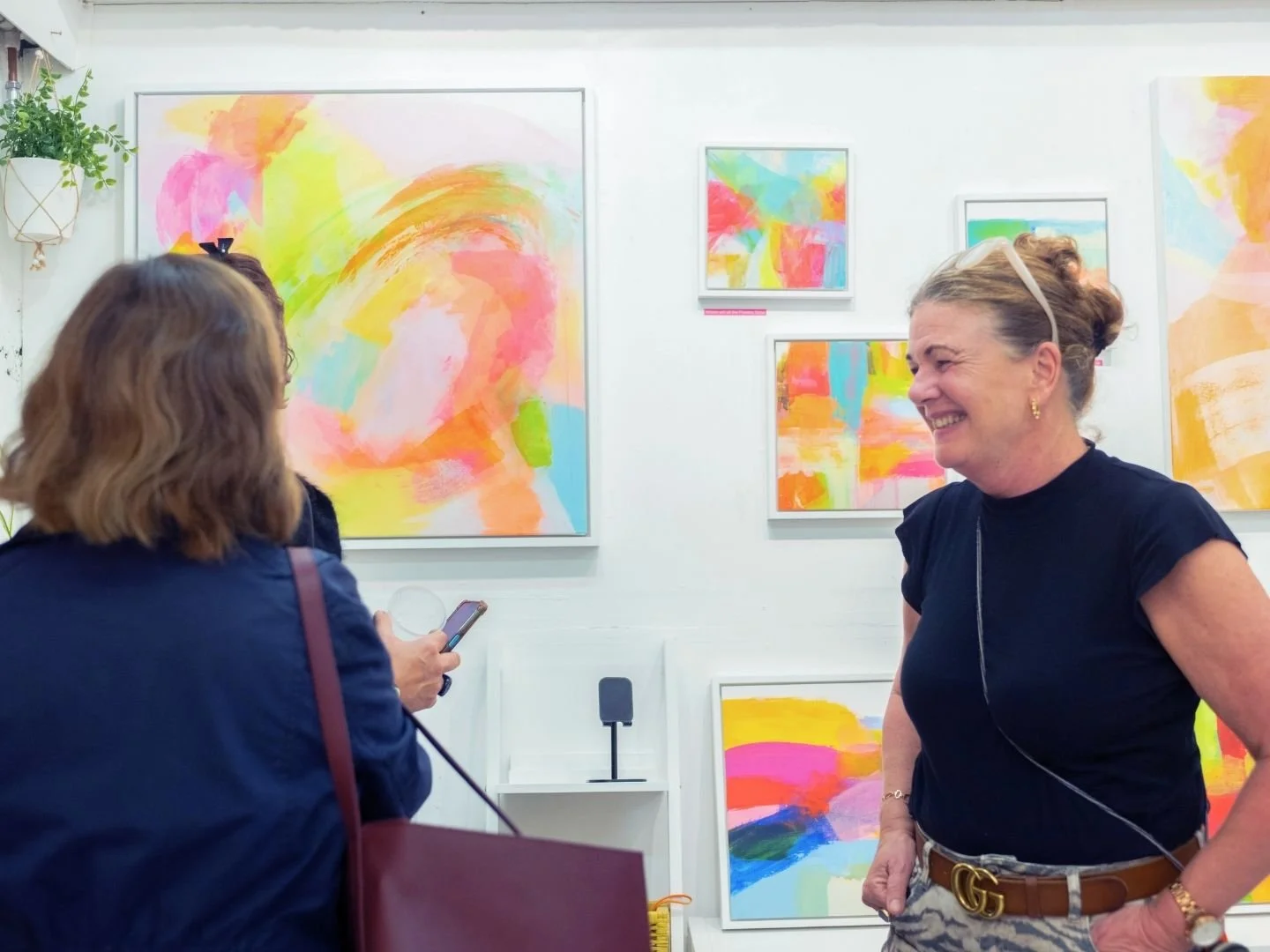 Two women are inside an art gallery, smiling and engaging in conversation. One woman, with brown hair, is facing away from the camera, holding a smartphone. The other woman, with blonde hair tied back and wearing glasses on her head, is visible smiling broadly. The gallery walls display colorful abstract paintings in various sizes.