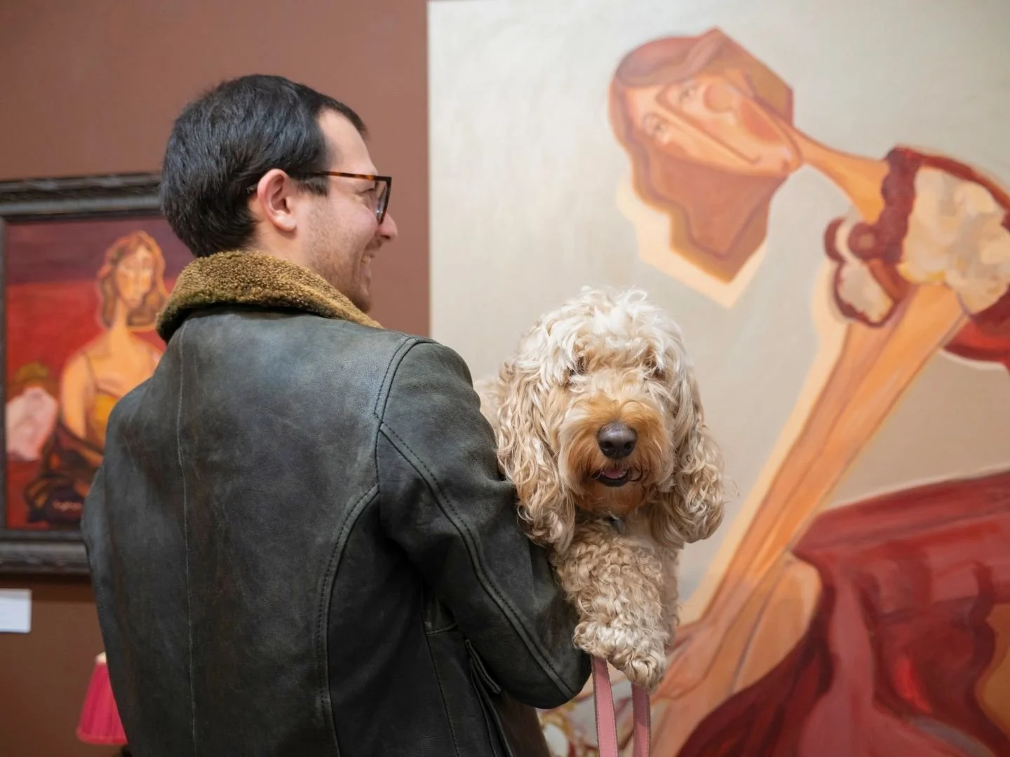 A man smiling while holding a curly-haired dog in an art gallery, with paintings in the background.
