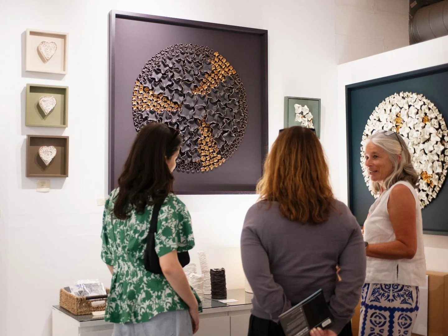 Three women conversing in an art gallery with colorful framed artwork on the white wall behind them, including a large circular piece made of small dark and gold elements.