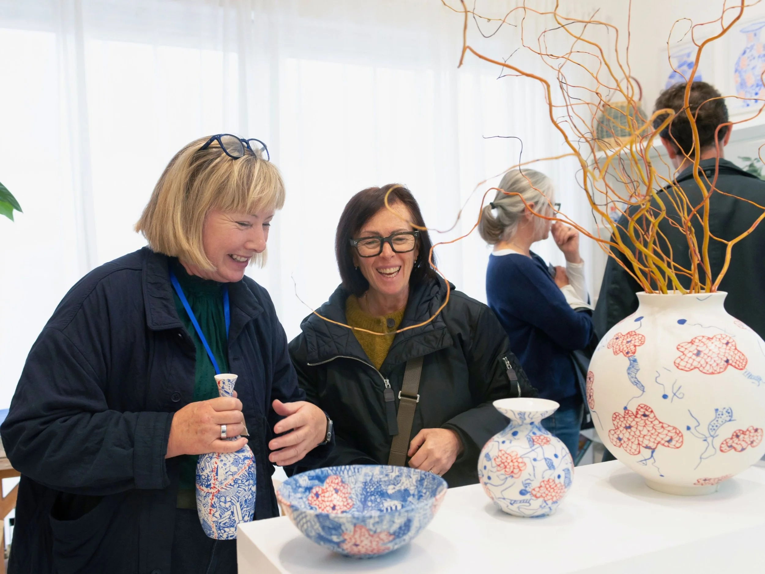 A ceramicist is holding one of her porcelain vases, showing her original artworks to a visitor from an open art studios event. The visitor and the artist are smiling.