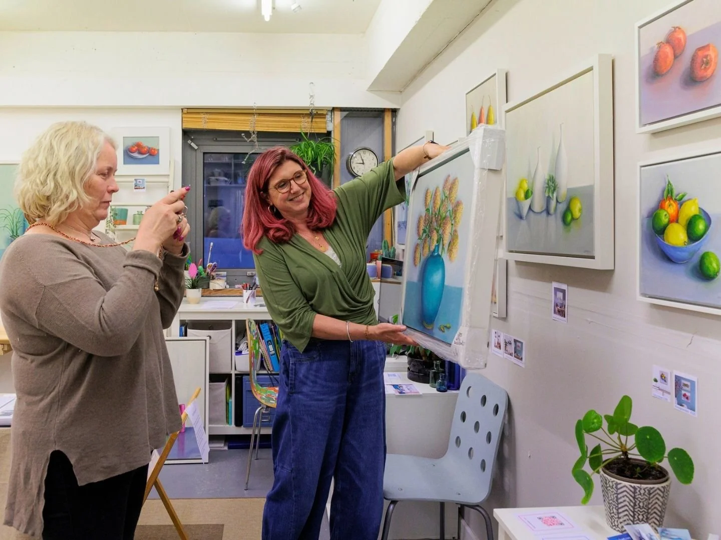 Two women viewing a painted still life of a blue vase with orange flowers, displayed on an easel in an art studio. The artist has framed fruit paintings on the wall and various art supplies and plants around.