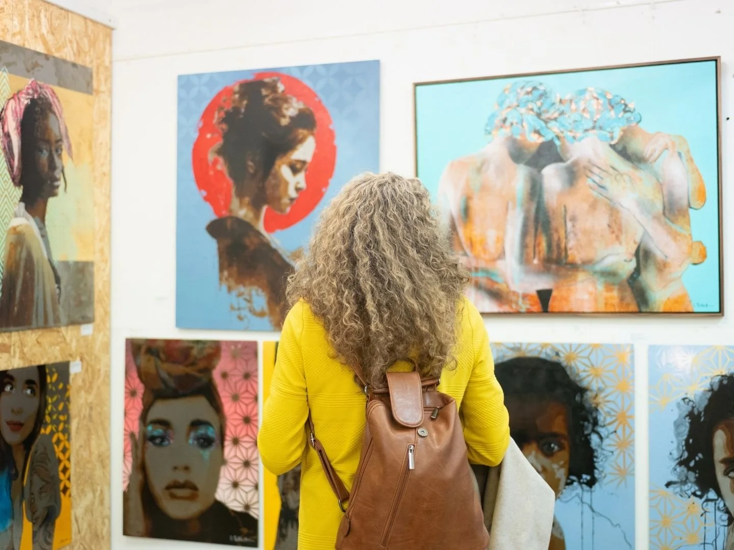 A woman with curly blonde hair wearing a yellow jacket and brown backpack stands in an art studio, viewing colourful portraits and paintings of women on the wall.