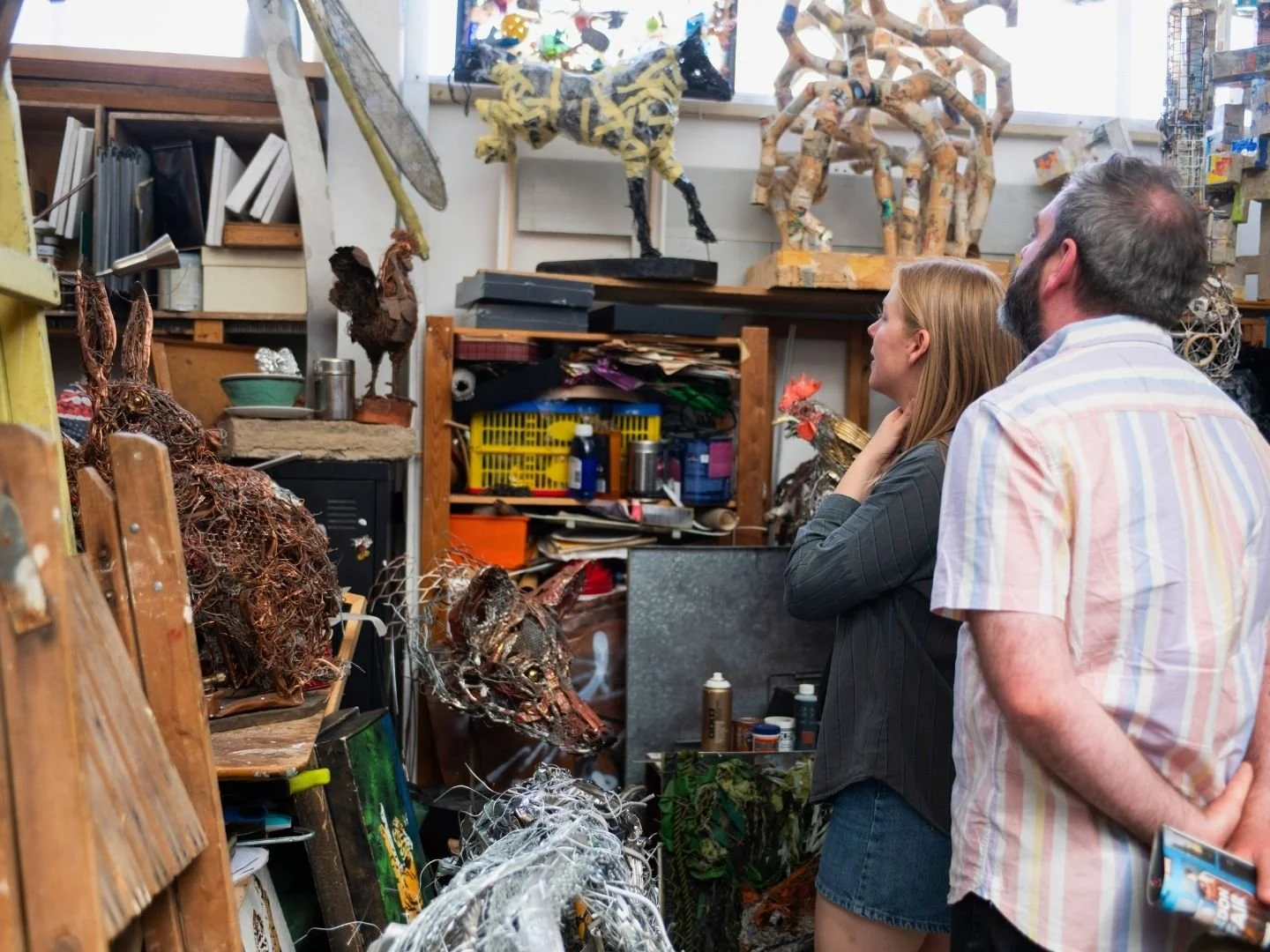 Two people, a man and a woman, are observing sculptures in an art studio filled with various art pieces, sculptures, and art supplies.