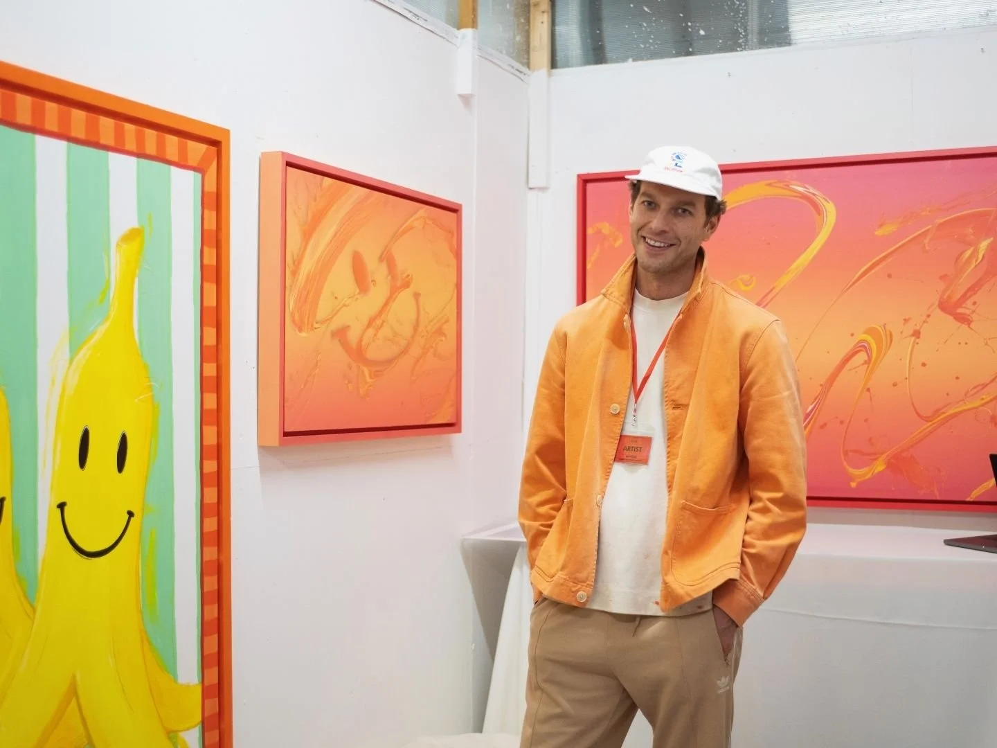 An artist smiling at his art studio, standing next to colourful abstract paintings with bright orange, yellow, and pink hues, wearing a white cap, orange jacket, and beige pants.